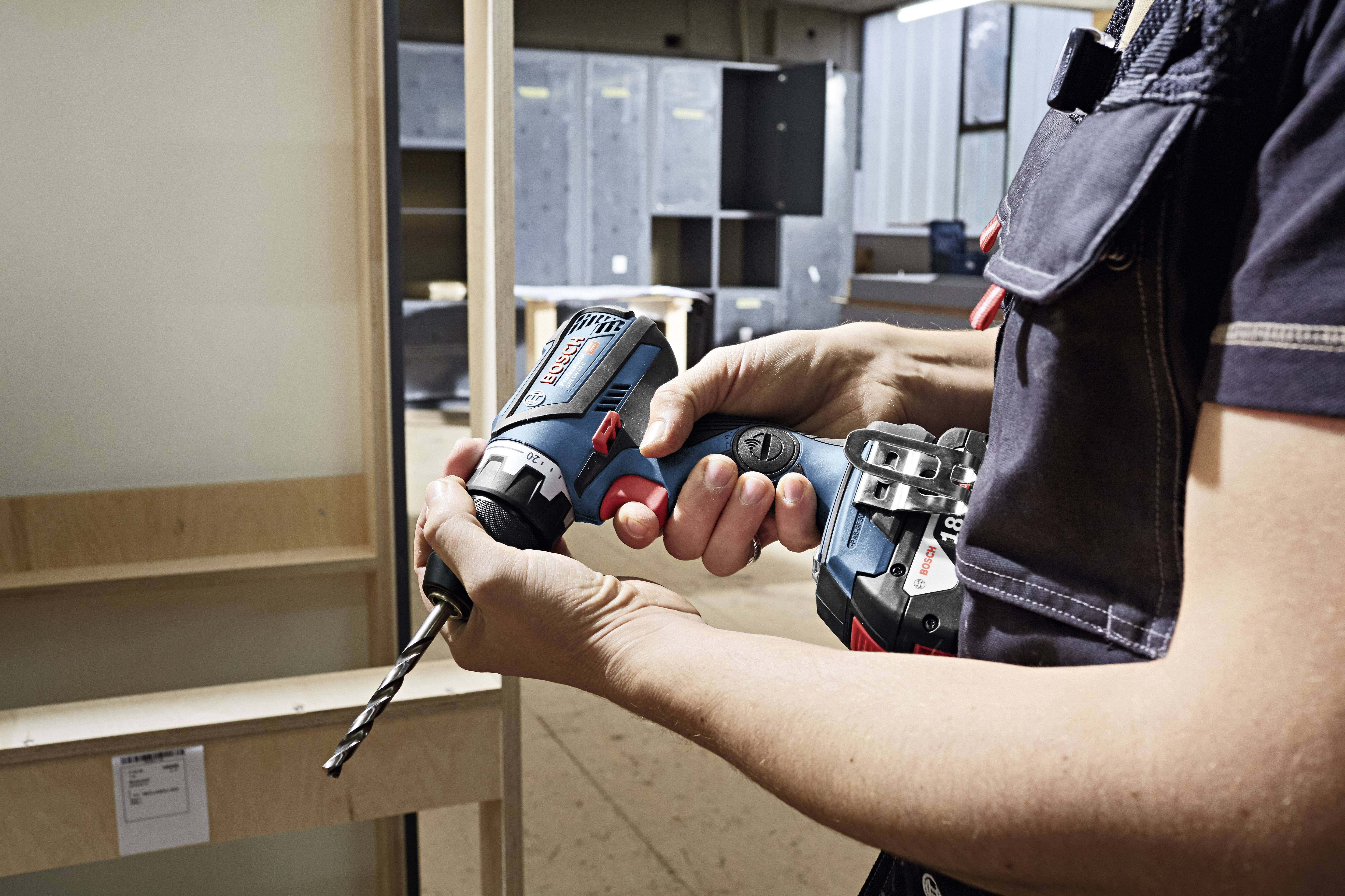 Eine Person hält einen blauen Akkubohrschrauber mit einem eingesetzten Bohrer in einer Werkstatt, bereit zu bohren. Im Hintergrund sind Regale sichtbar.