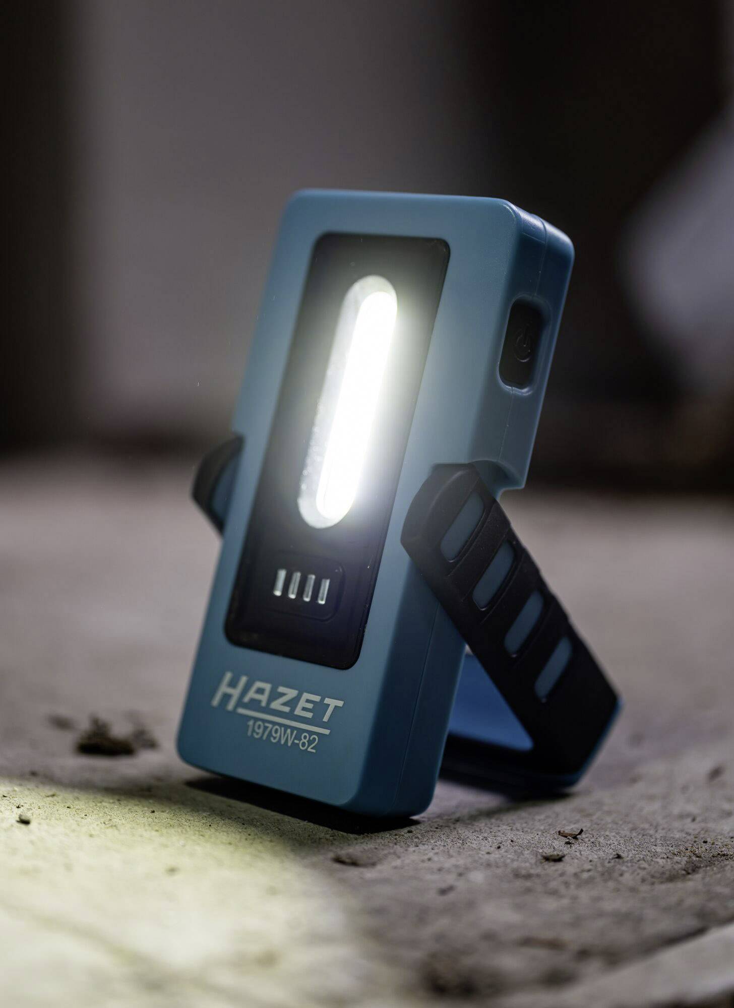 Tragbare, blaue LED-Arbeitsleuchte auf einem Boden, erstrahlt hell. Text auf dem Licht: 'Hazet 1979W-82'.