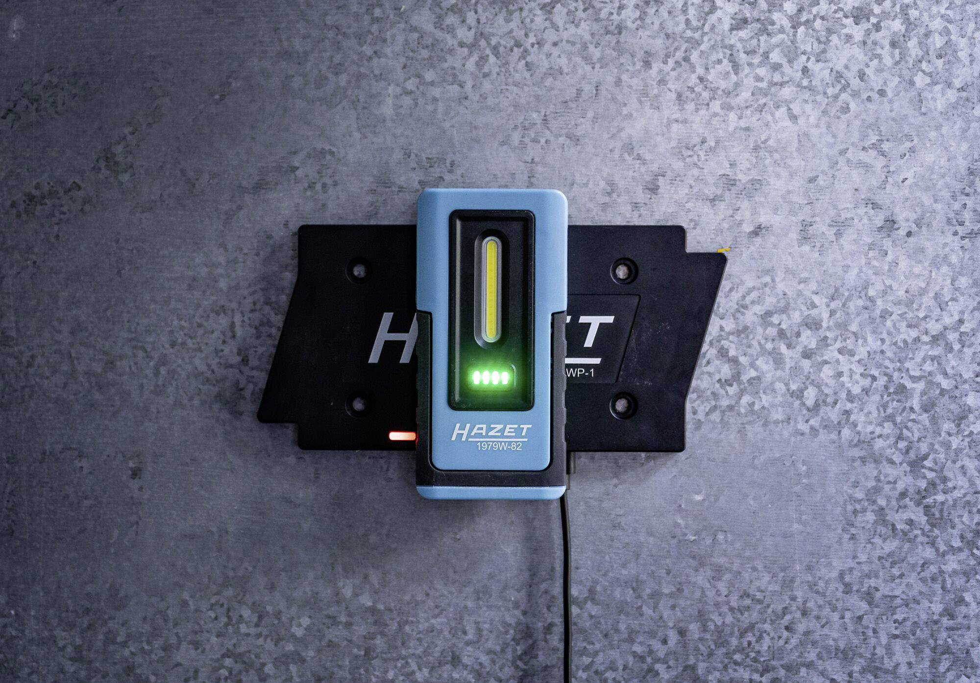 Gerät zur Wandmontage mit LED-Anzeige, zeigt 'HAZET' an. Es wird über ein Kabel mit Strom versorgt, auf metallischer Oberfläche befestigt.