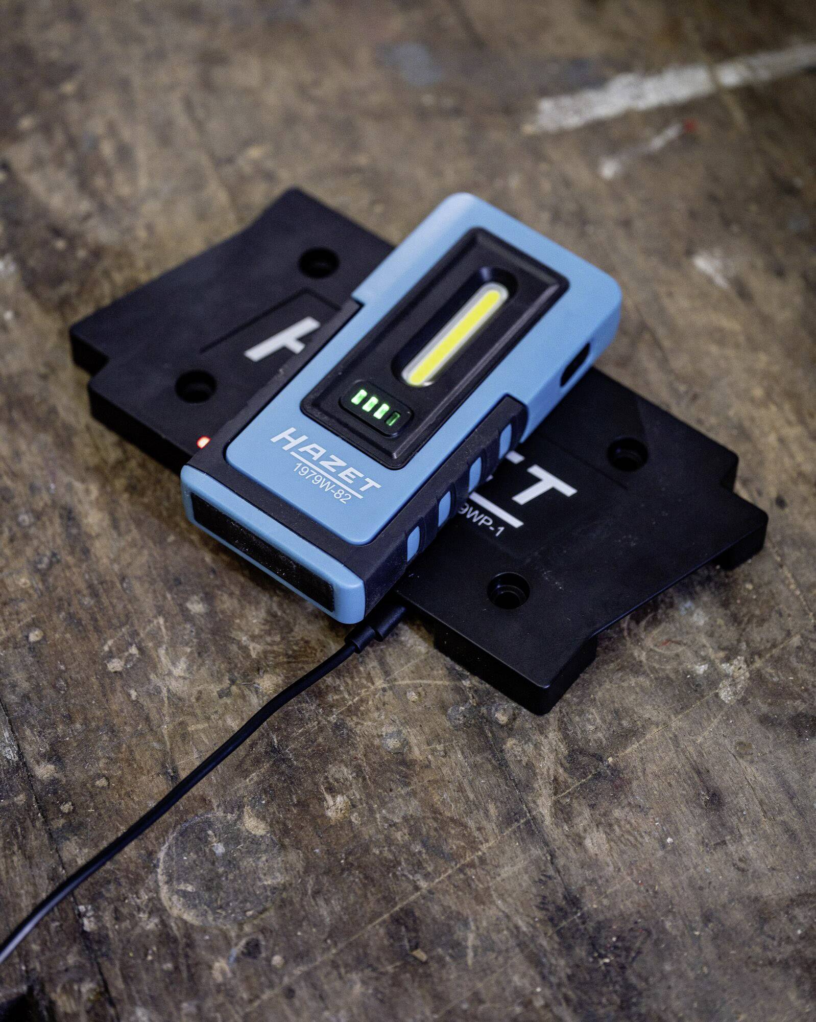 Eine blaue LED-Arbeitslampe von Hazet wird auf einer schwarzen Oberfläche aufgeladen und zeigt einen Ladezustandsindikator an.