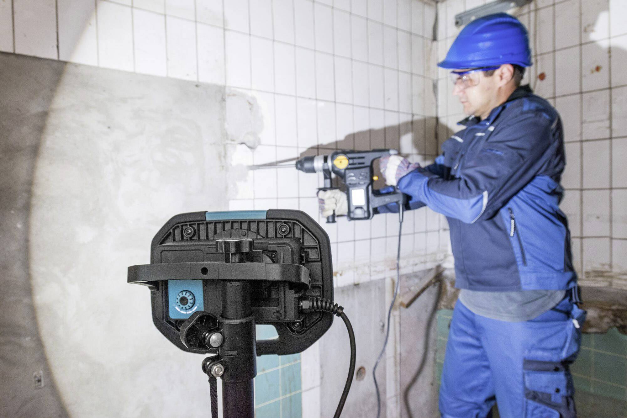 Ein Bauarbeiter in blauer Schutzkleidung und Helm arbeitet mit einer Bohrmaschine an einer gefliesten Wand in einem Badezimmer.