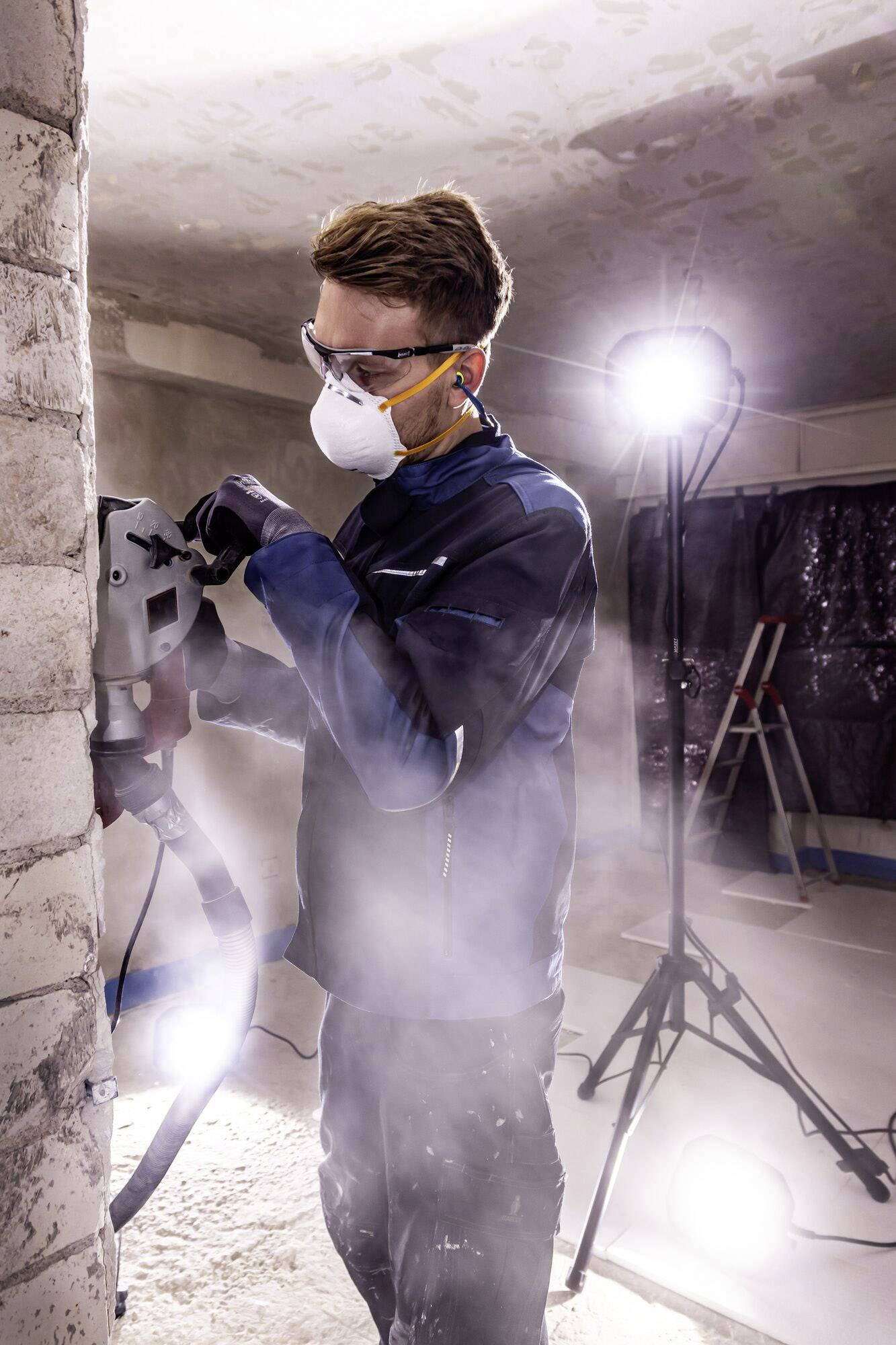 Arbeiter mit Atemmaske schleift Wand, beleuchtet von zwei starken Lampen. Schutzkleidung und Staub sichtbar, Hinweis auf Renovierungsarbeiten.