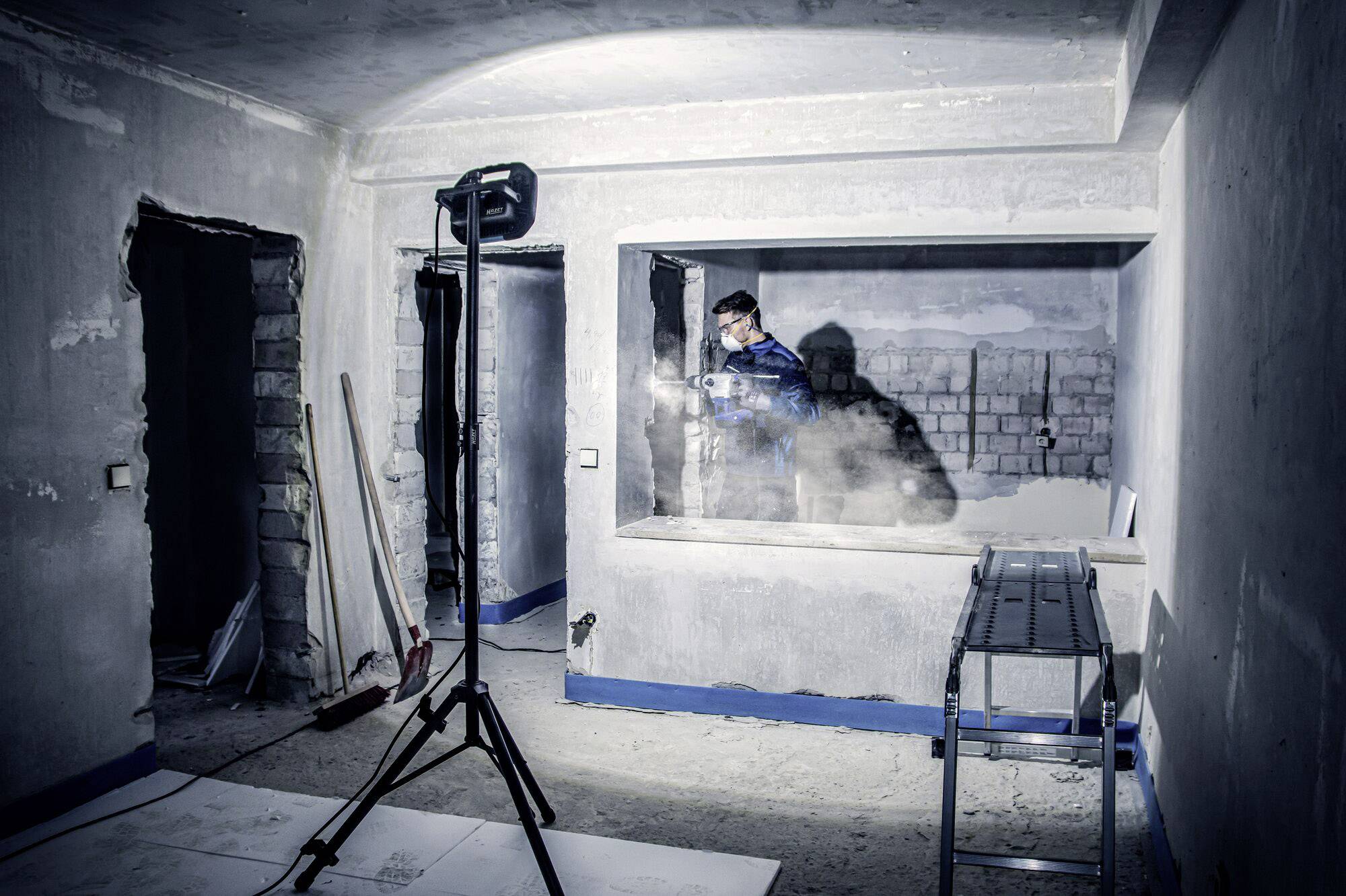 Eine Person arbeitet in einem unfertigen Raum mit Baustrahler. Der Raum ist im Rohbauzustand, Wände unverputzt, Bauwerkzeuge sichtbar.