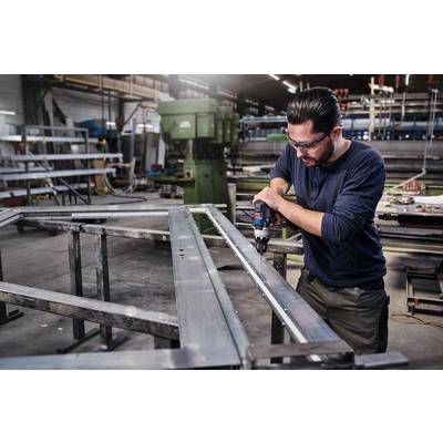 Ein Mann trägt Schutzbrille und bohrt ein Metallrahmen in einer Werkstatt. Im Hintergrund sind Maschinen und weitere Materialien zu sehen.