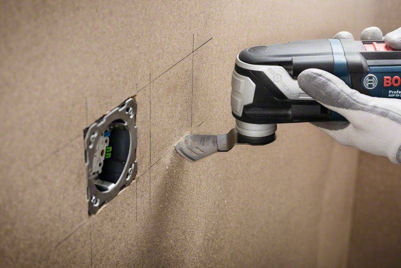 Eine Hand führt ein Multitool aus, das ein Loch in eine Trockenbauwand schneidet. Daneben ist eine Steckdosenöffnung zu sehen.