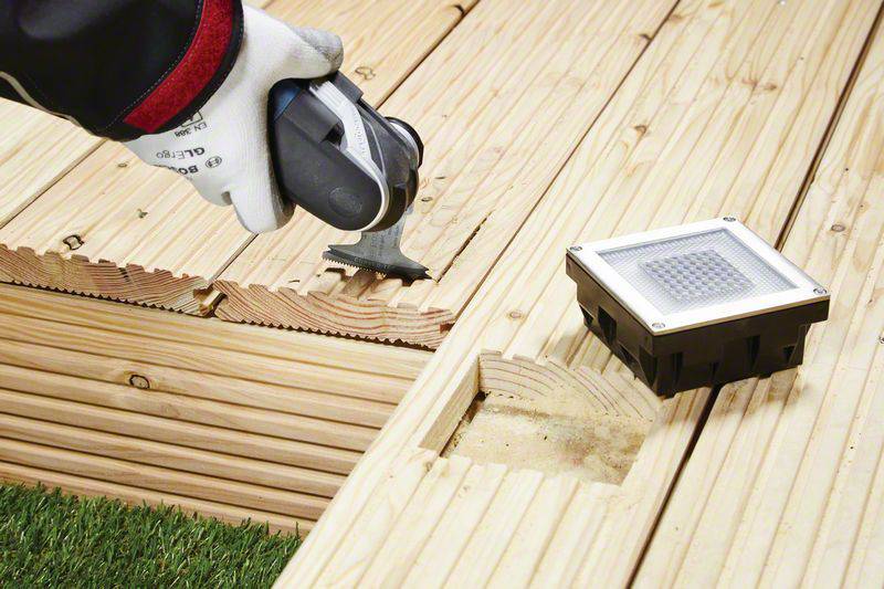 Arbeiter in Handschuhen installiert ein Holzdeck und verwendet Werkzeug für Präzision. Ein Bodenlicht wird in das Deck integriert.