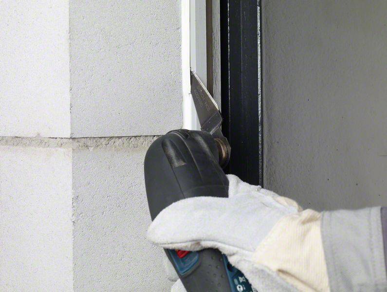 Eine Person trägt Handschuhe und verwendet ein Elektrowerkzeug, um eine Fuge zwischen einer weißen Mauer und einem Fensterrahmen zu entfernen.