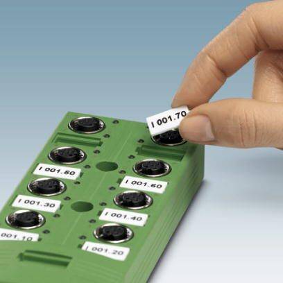 Hand wechselt Beschriftungsschild an einem grünen, elektronischen Gerät mit runden Steckplätzen, nummeriert von '001.10' bis '001.70'.