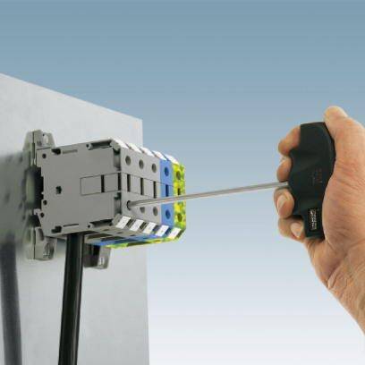 Eine Hand hält einen Schraubendreher und zieht eine Schraube an einem elektrischen Schaltgerät an einer grauen Wand fest.
