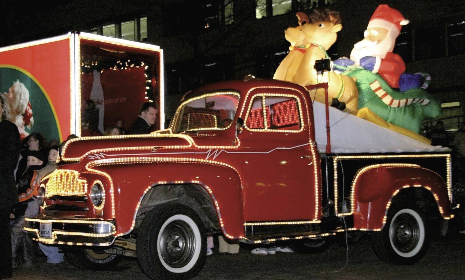 Ein roter Lastwagen mit Lichterketten transportiert Weihnachtsdekoration, darunter ein großer aufblasbarer Weihnachtsmann und ein Rentier.