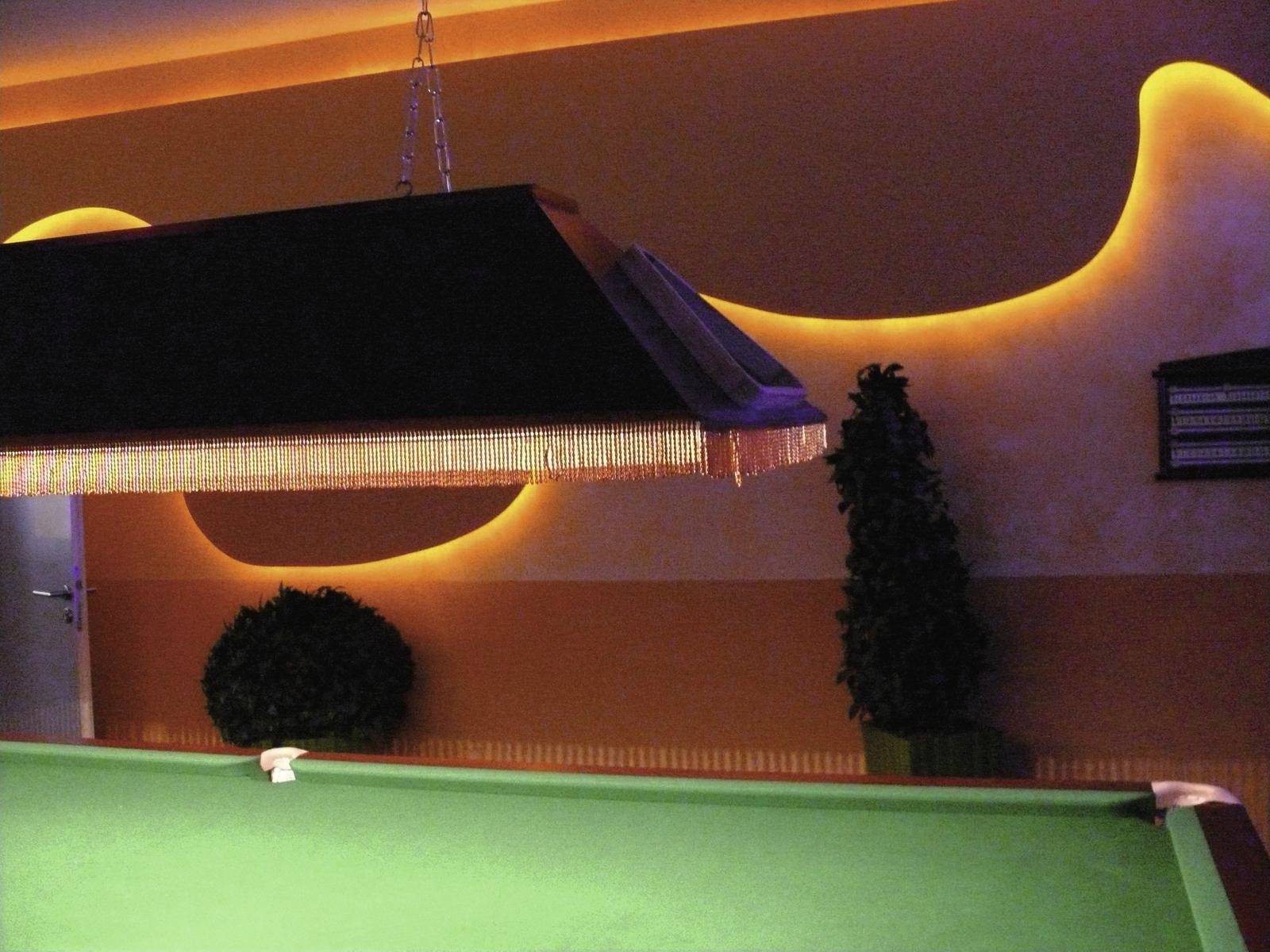 Ein Billardtisch in einem stimmungsvoll beleuchteten Raum mit dekorativen Wänden und Pflanzen. Eine hängende Lampe beleuchtet den Tisch.