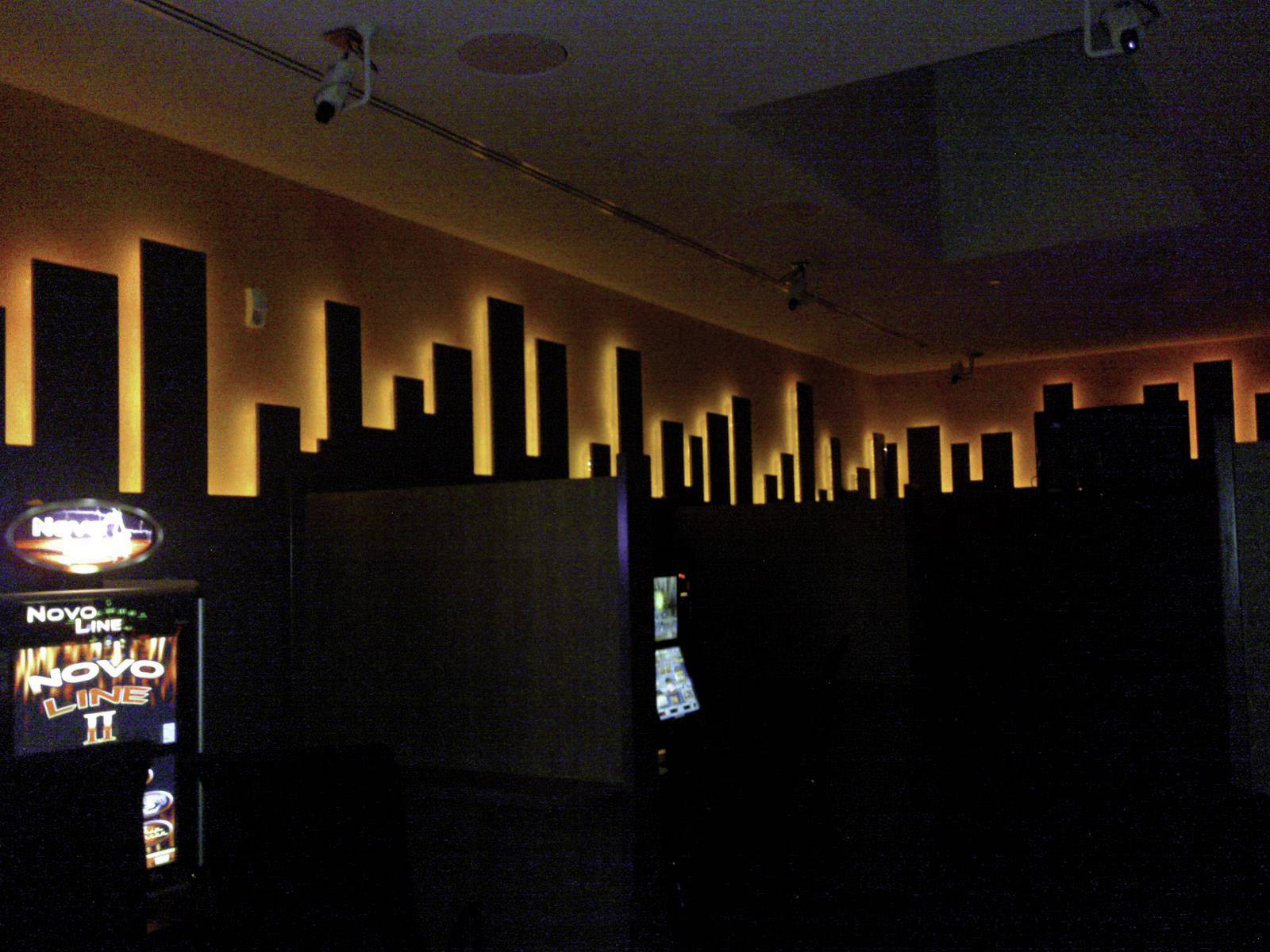 Eine dunkle Spielehalle mit leuchtenden Spielautomaten. Die Wand dahinter zeigt eine stilisierte Skyline mit beleuchteten Akzenten.