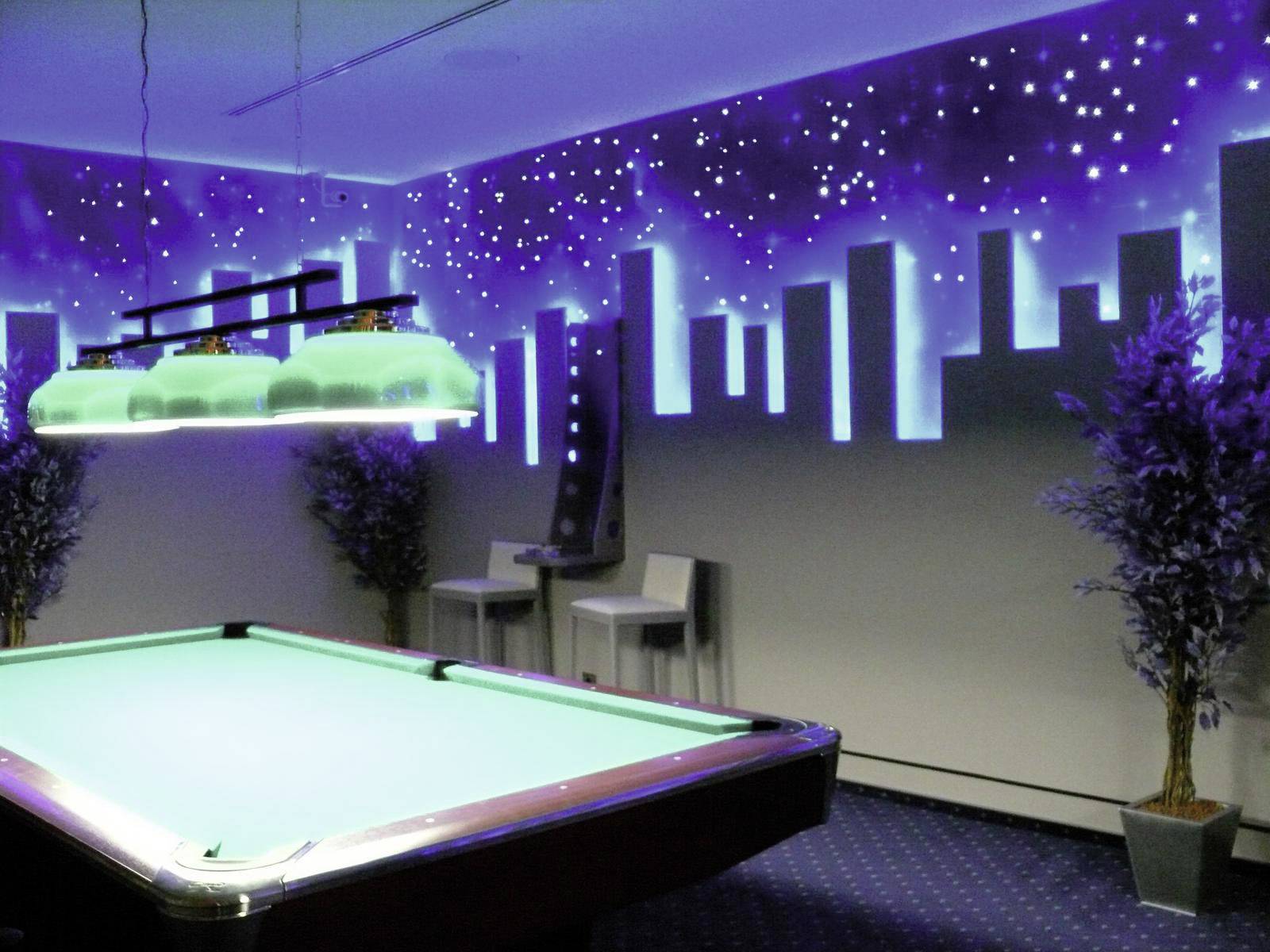 Billardzimmer mit beleuchtetem Tisch, bemalter Skyline an der Wand und dekorativen Pflanzen. Stimmungsvolles Licht schafft urbane Atmosphäre.
