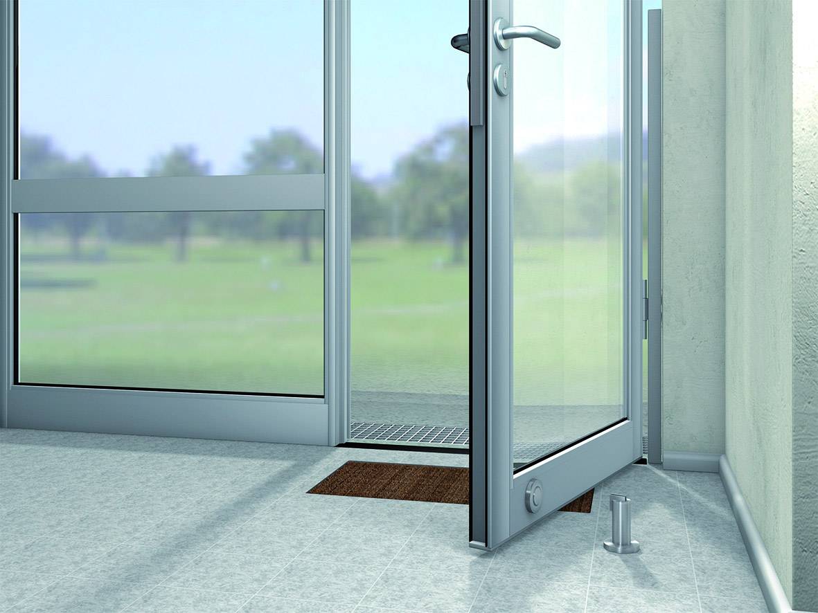 Eine Glastür ist leicht geöffnet und führt nach draußen zu einer grünen Wiese. Ein kleiner brauner Teppich liegt vor der Tür.