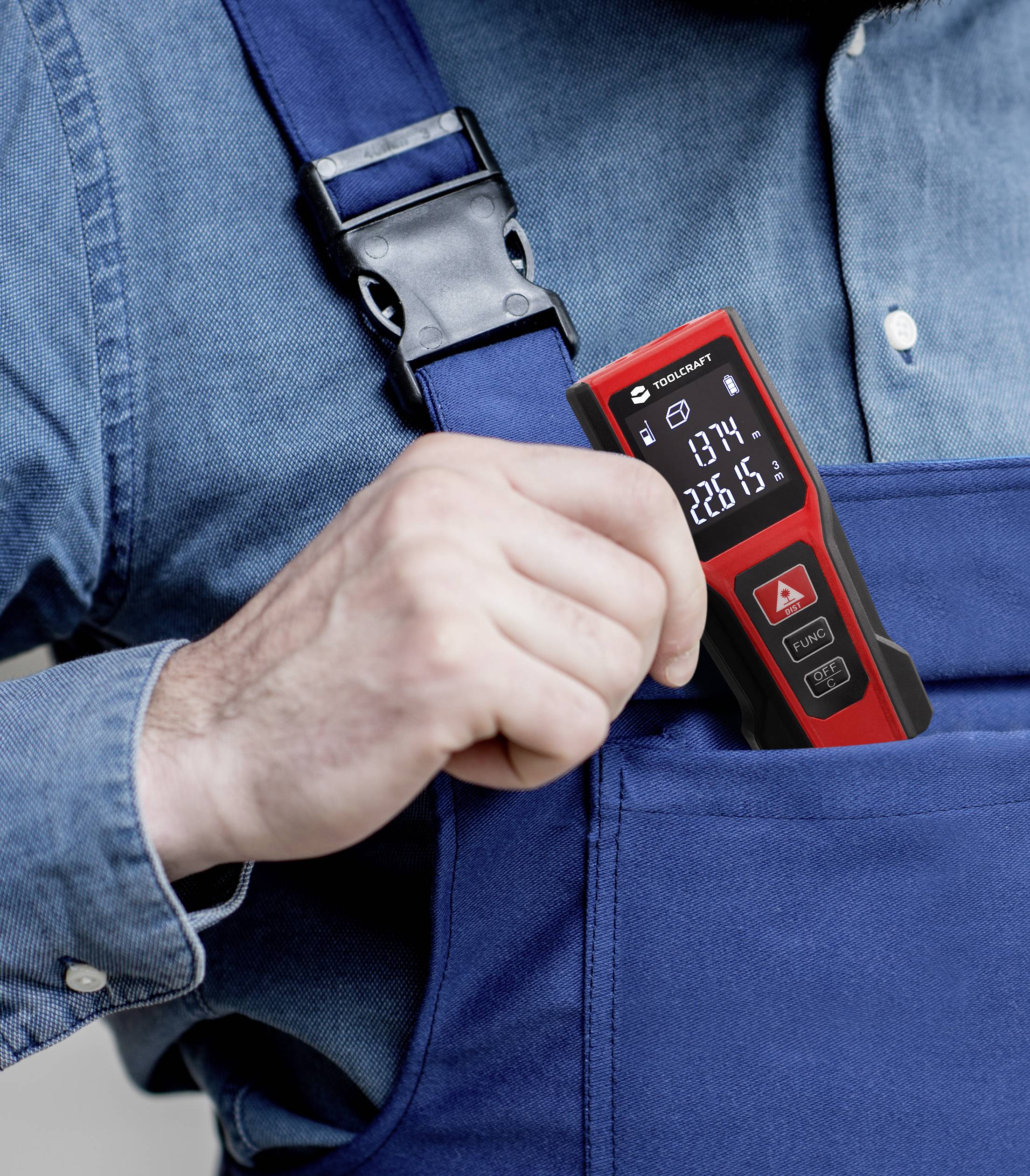 Person in blauer Latzhose steckt ein rotes elektronisches Messgerät in die Brusttasche. Das Display zeigt Zahlenwerte an.