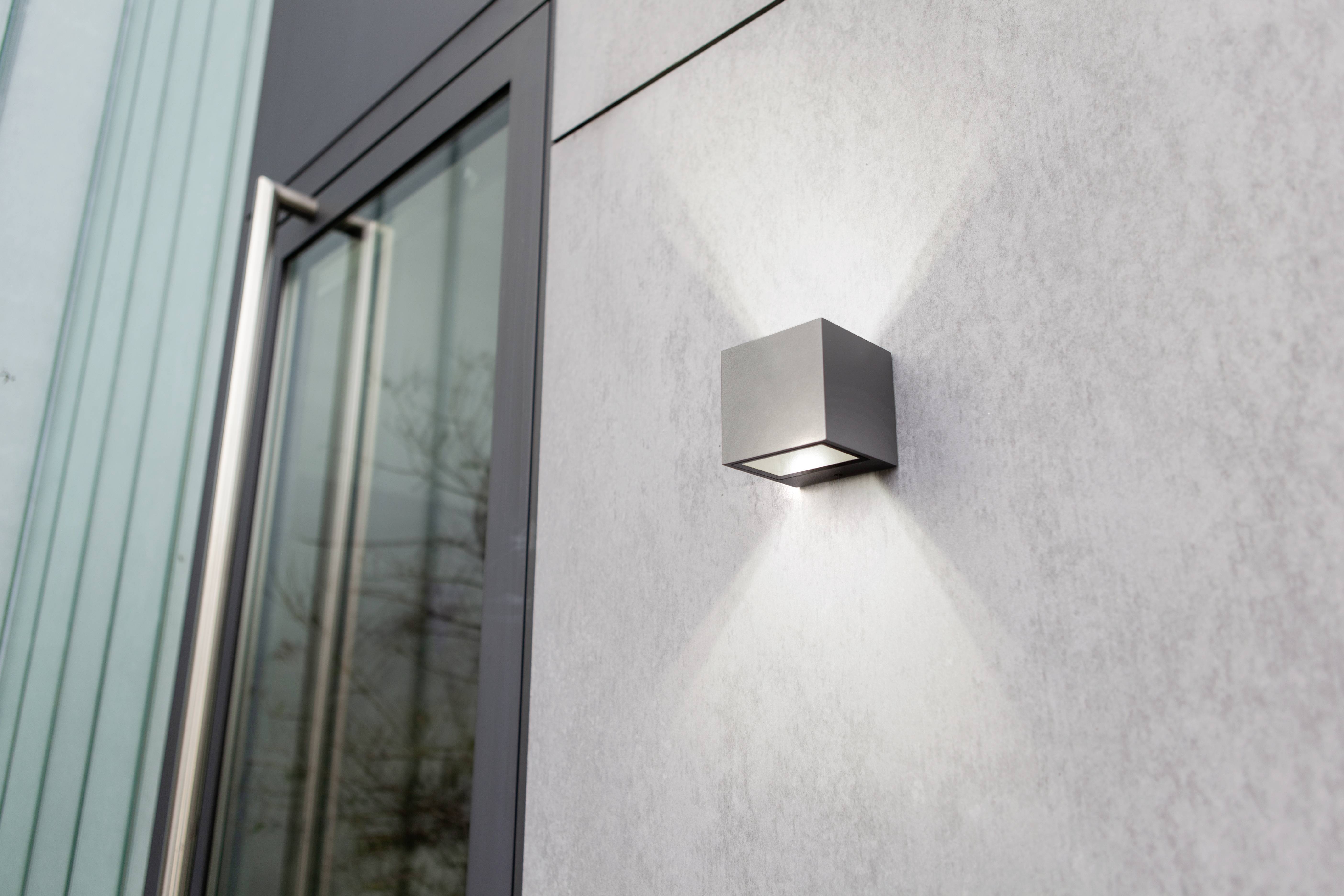 Außenleuchte in modernem, kubischem Design an grauer Wand, leuchtet nach oben und unten. Fenster im Hintergrund.