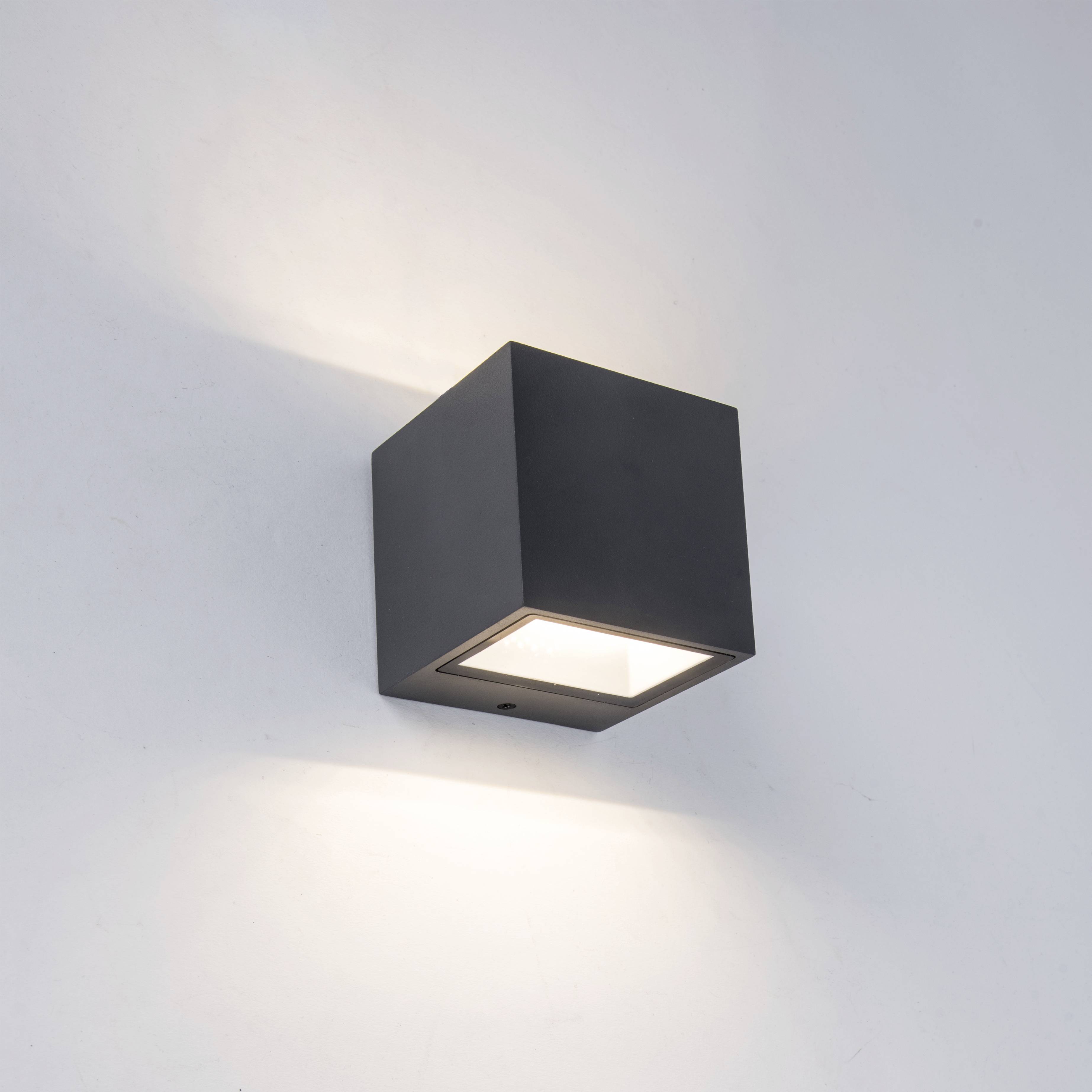 Schwarze würfelförmige Wandlampe, die nach oben und unten Lichtstrahlen auf eine weiße Wand wirft.