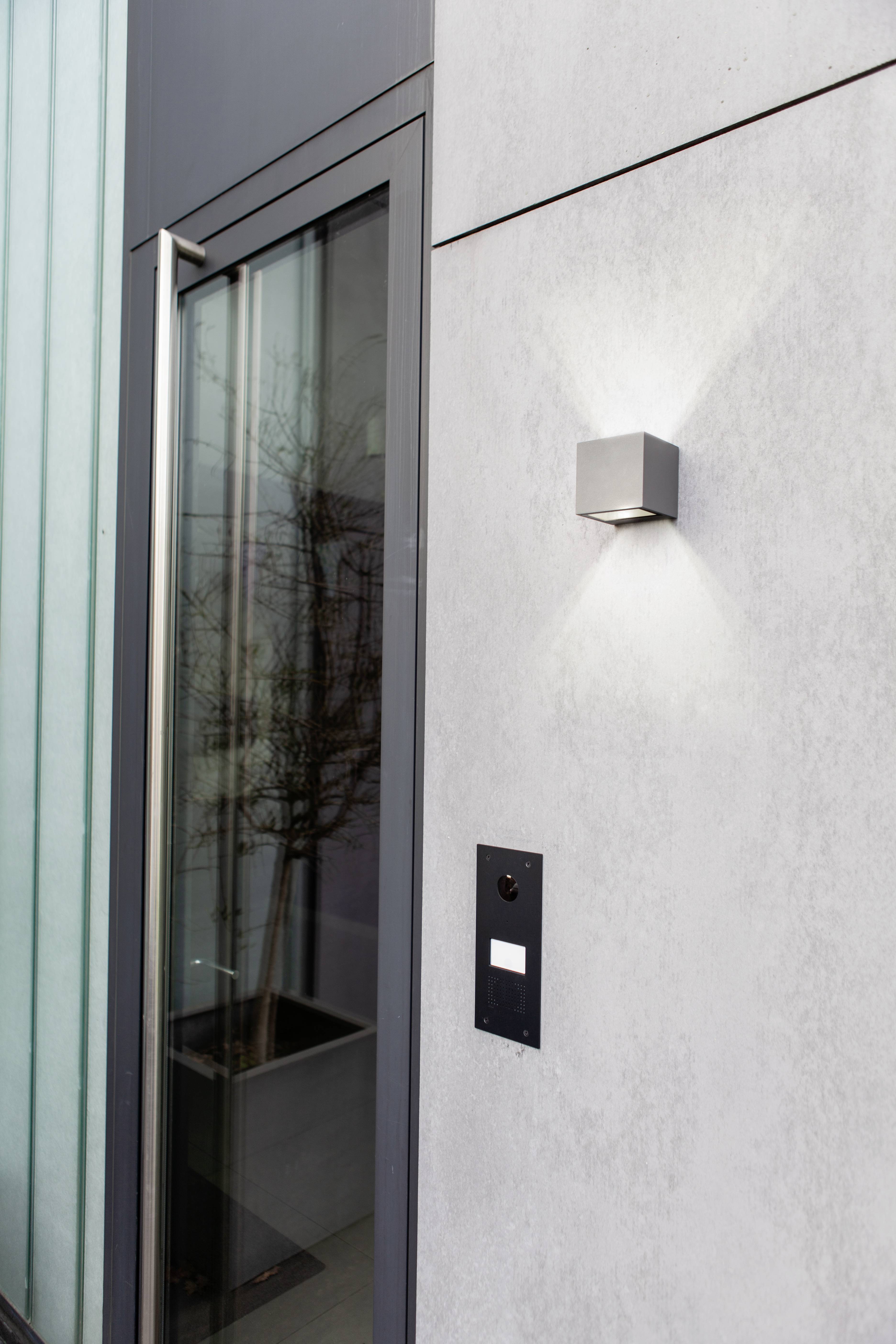 Eine moderne Außentür neben einer Betonwand mit einer quadratischen Wandleuchte, die ein weiches Licht verbreitet.