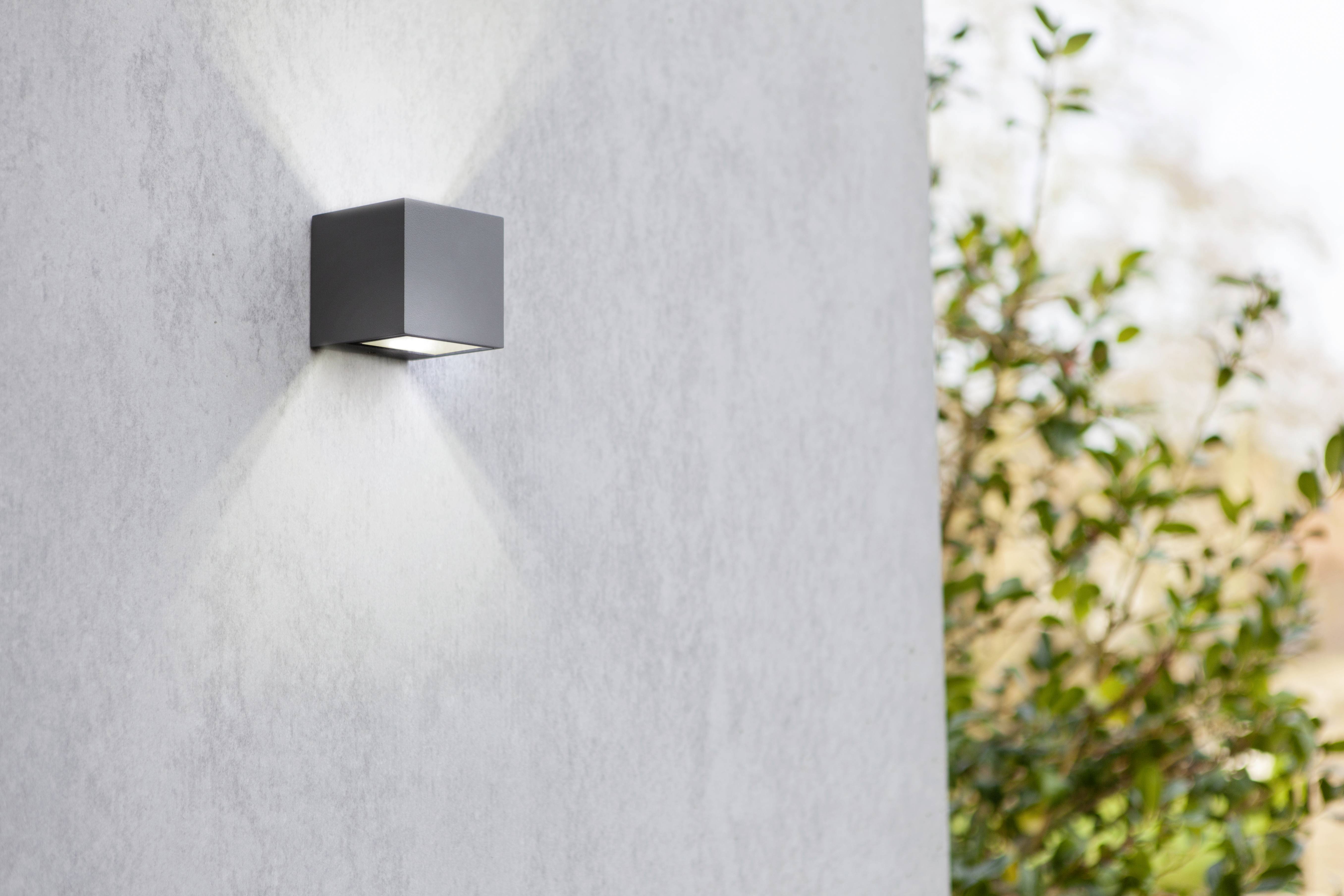 Eine moderne, graue Wandleuchte auf einer hellen Betonwand. Die Lampe strahlt Licht nach oben und unten aus. Rechts im Hintergrund sind unscharfe grüne Blätter sichtbar.