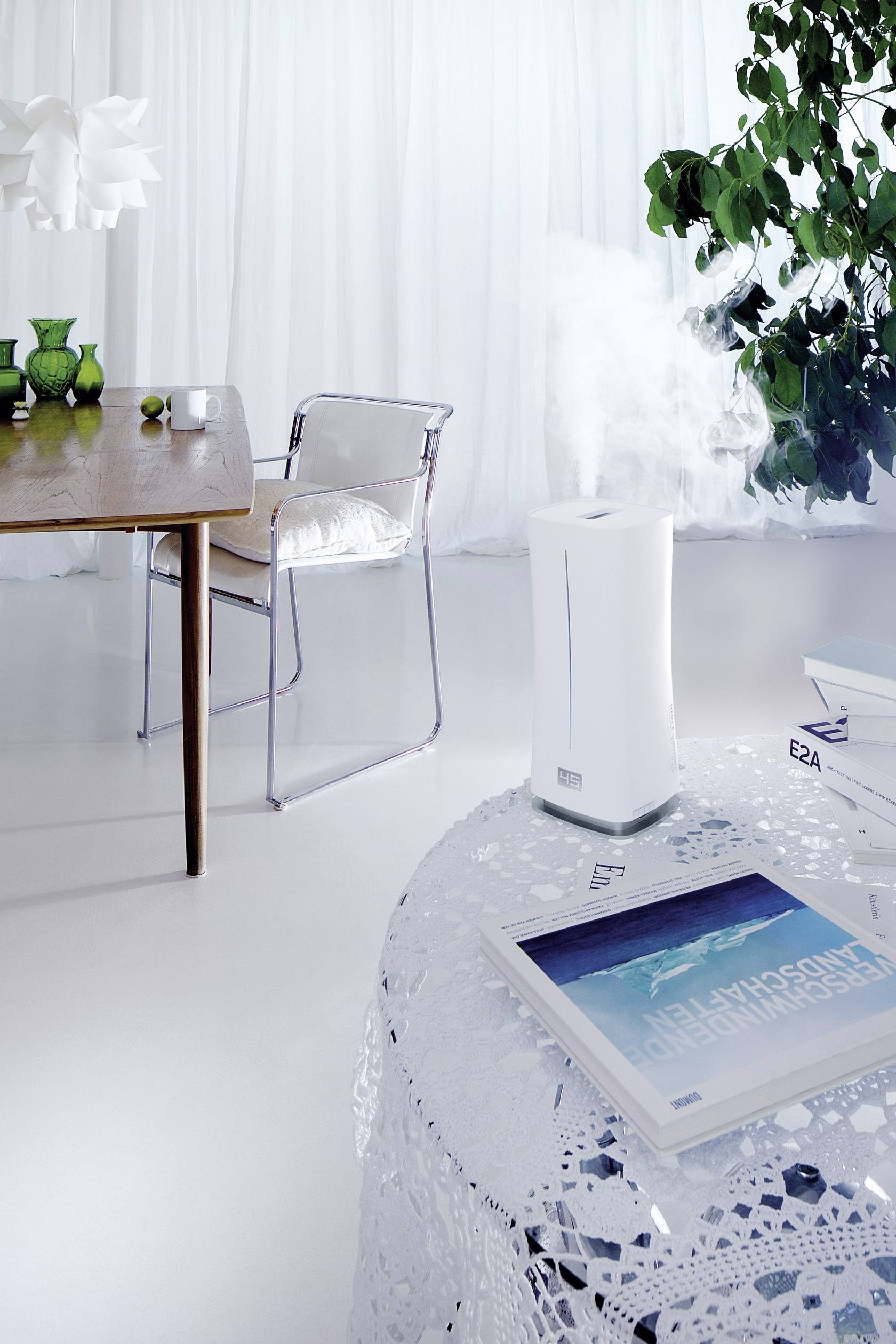 Moderner, heller Raum mit Tisch und Stühlen, Luftbefeuchter auf Tisch mit Wasserdampf, Pflanzen im Hintergrund, helle Vorhänge.