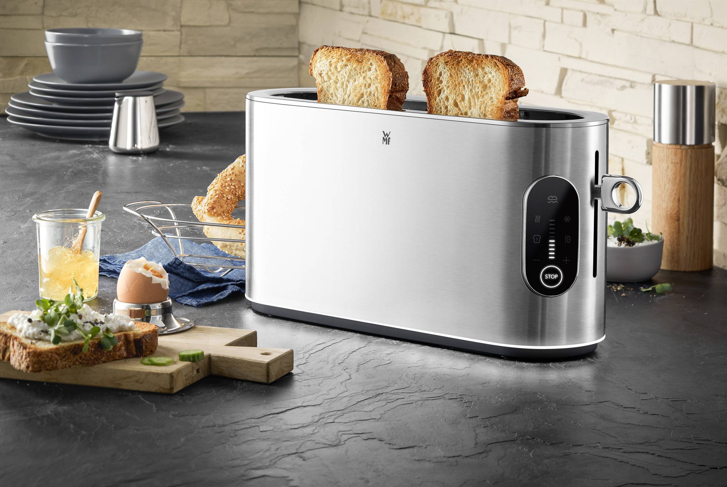 Ein Edelstahl-Toaster mit zwei Scheiben geröstetem Brot in einer modernen Küche mit Frühstücksleckereien und Geschirr im Hintergrund.