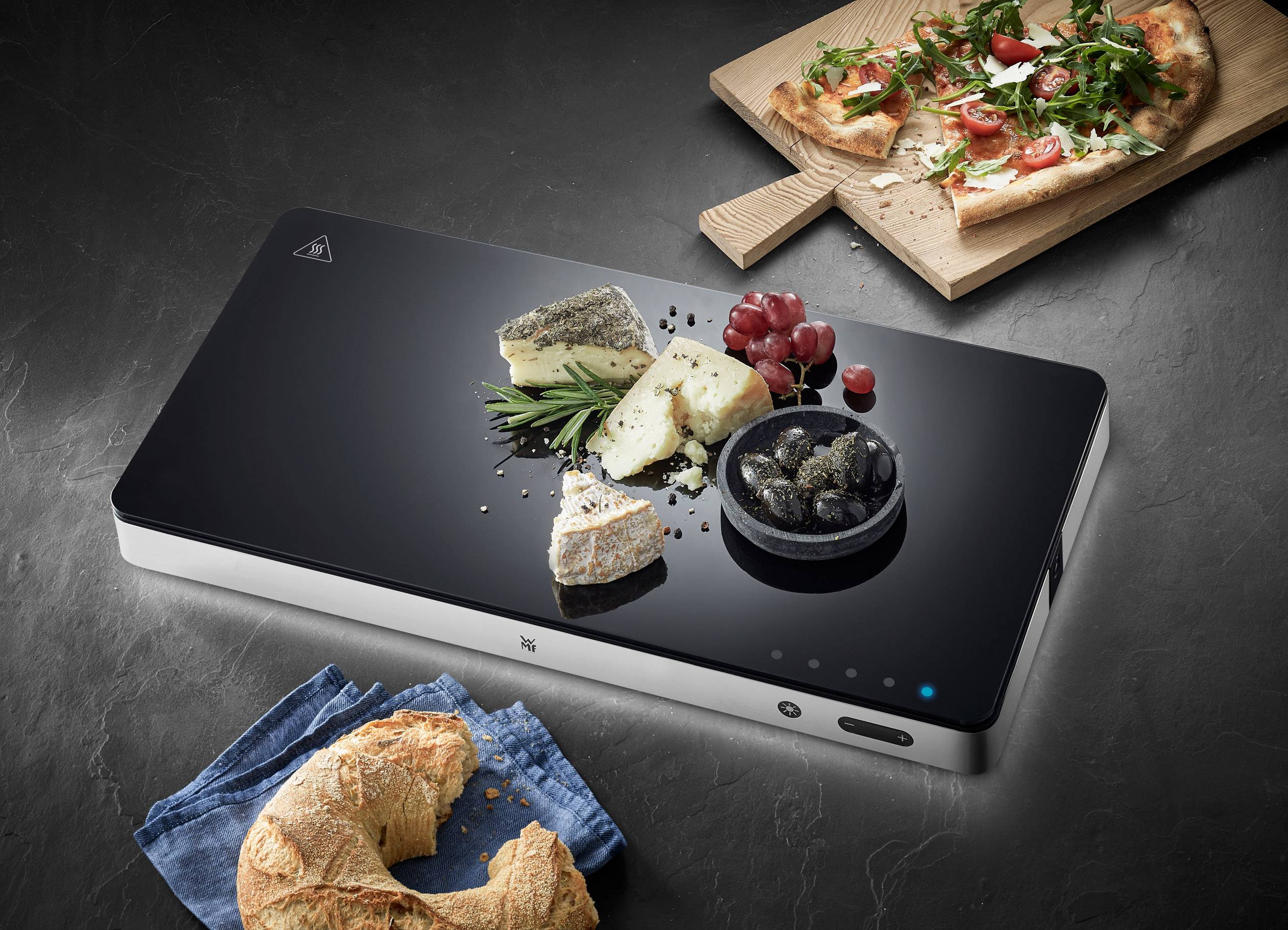 Ein elegantes schwarzes Induktionskochfeld mit verschiedenen Käsesorten, Trauben und Oliven darauf. Im Hintergrund eine Pizza und ein Croissant.