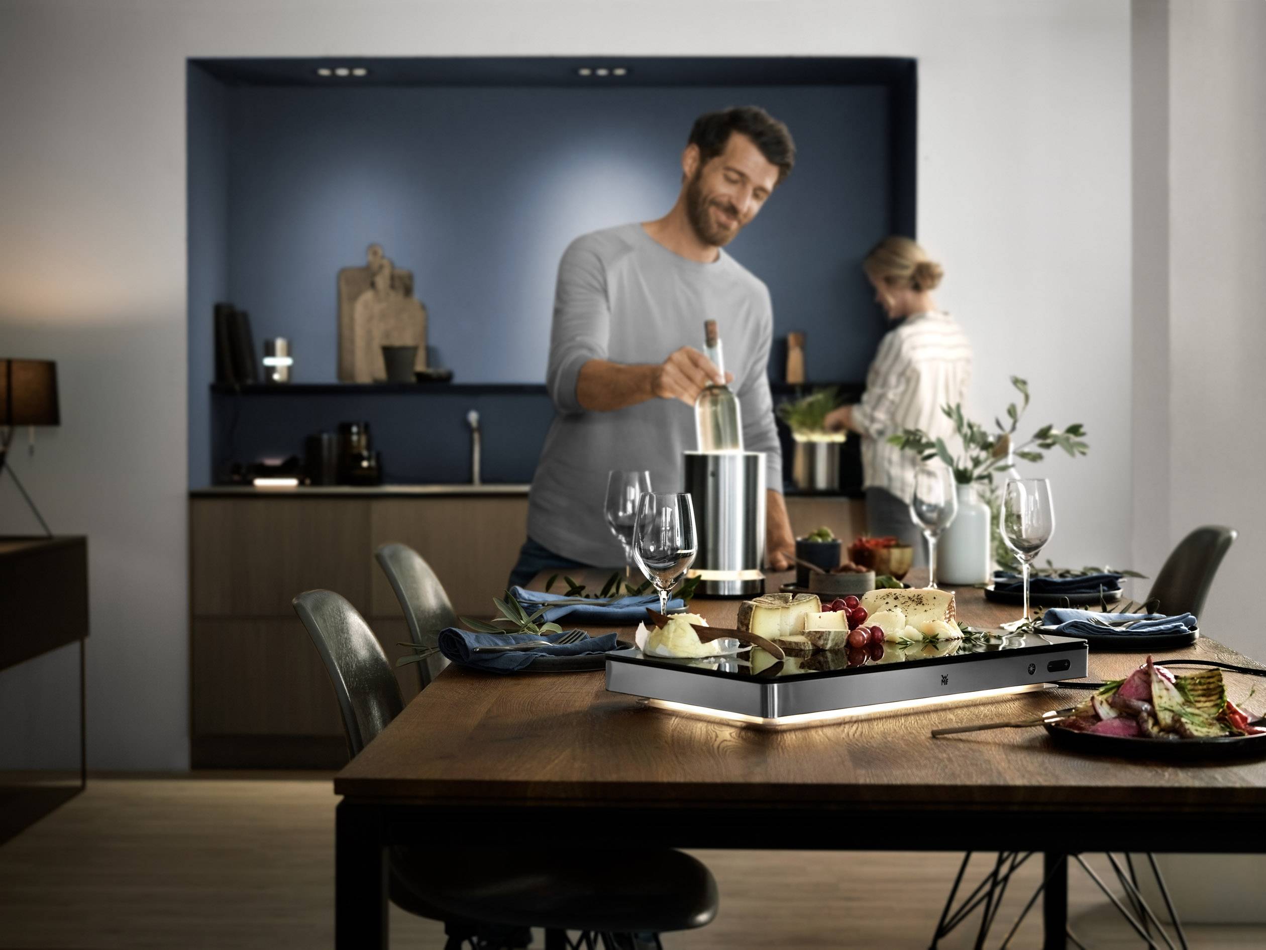 Ein Mann bereitet Essen am Tisch vor, während eine Person im Hintergrund in der Küche kocht. Der Raum wirkt modern und gemütlich.
