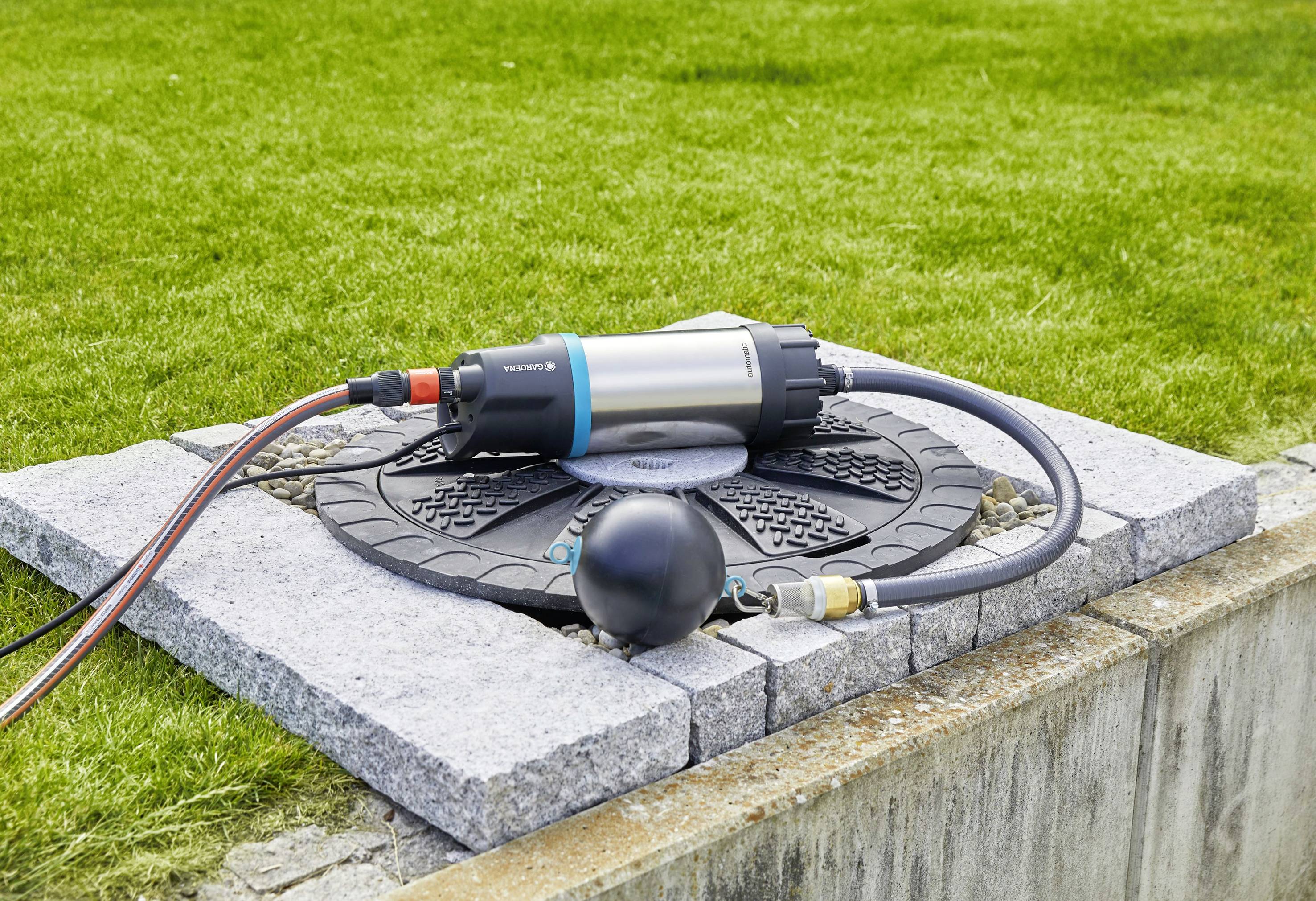 'Eine Gartenpumpe und Schlauch auf einem Kanaldeckel in einem Rasenbereich.'