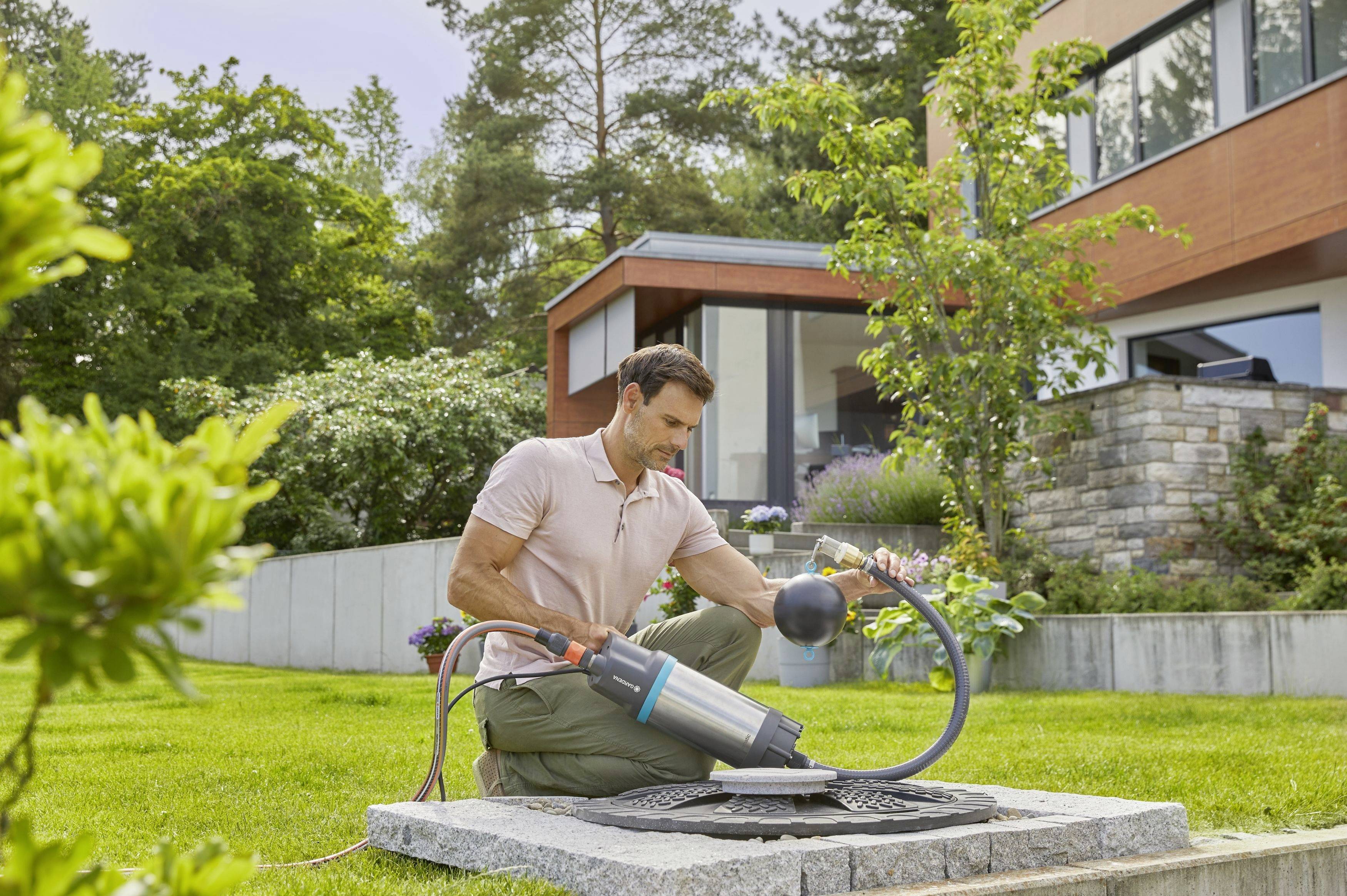 Ein Mann kniet im Garten und installiert eine Wasserpumpe an einem Abflussdeckel. Im Hintergrund ist ein modernes Haus zu sehen.