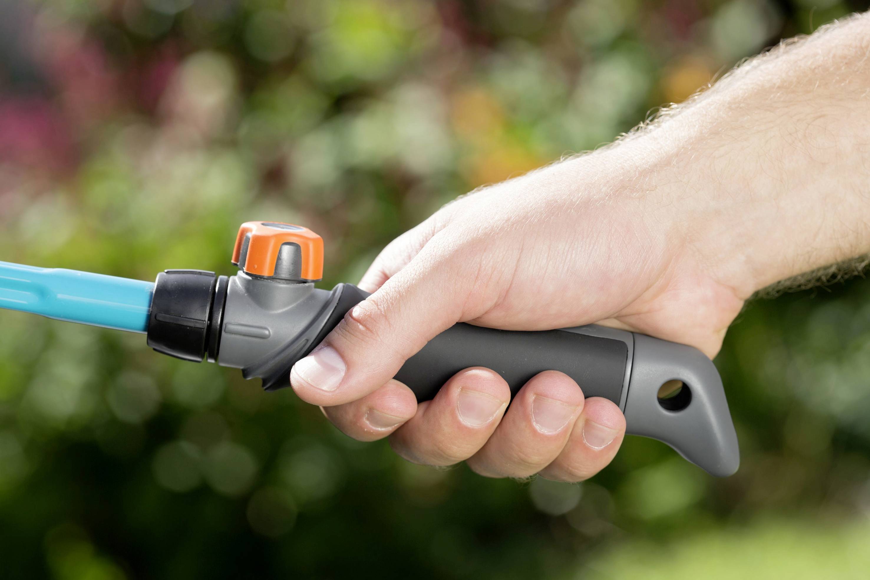 Eine Hand hält einen Gartenschlauch mit einem grauen Griff und einem orangefarbenen Ventil vor einem verschwommenen Hintergrund.
