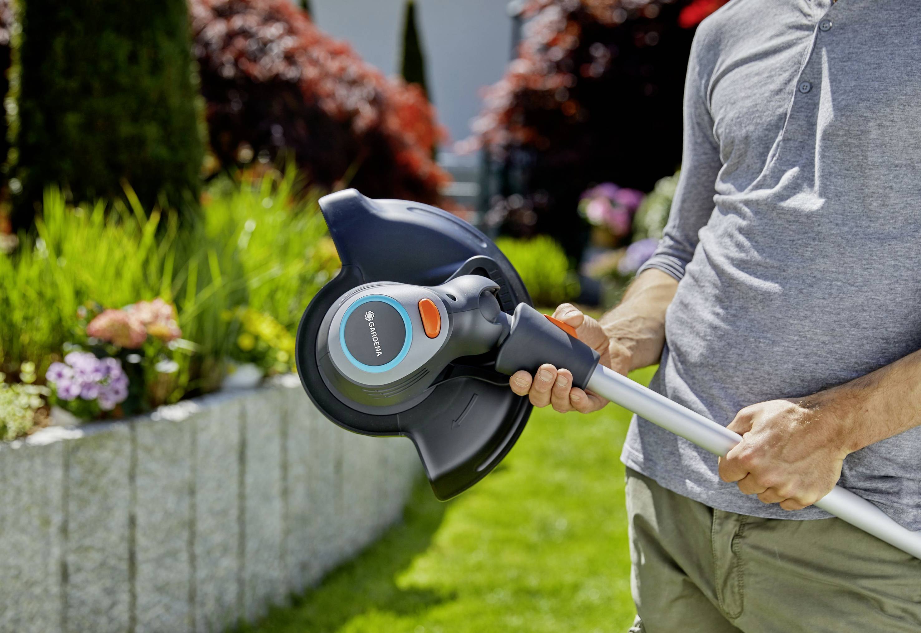 Person arbeitet im Garten und hält einen elektrischen Rasentrimmer. Im Hintergrund sind Blumenbeete und grüne Pflanzen zu sehen.
