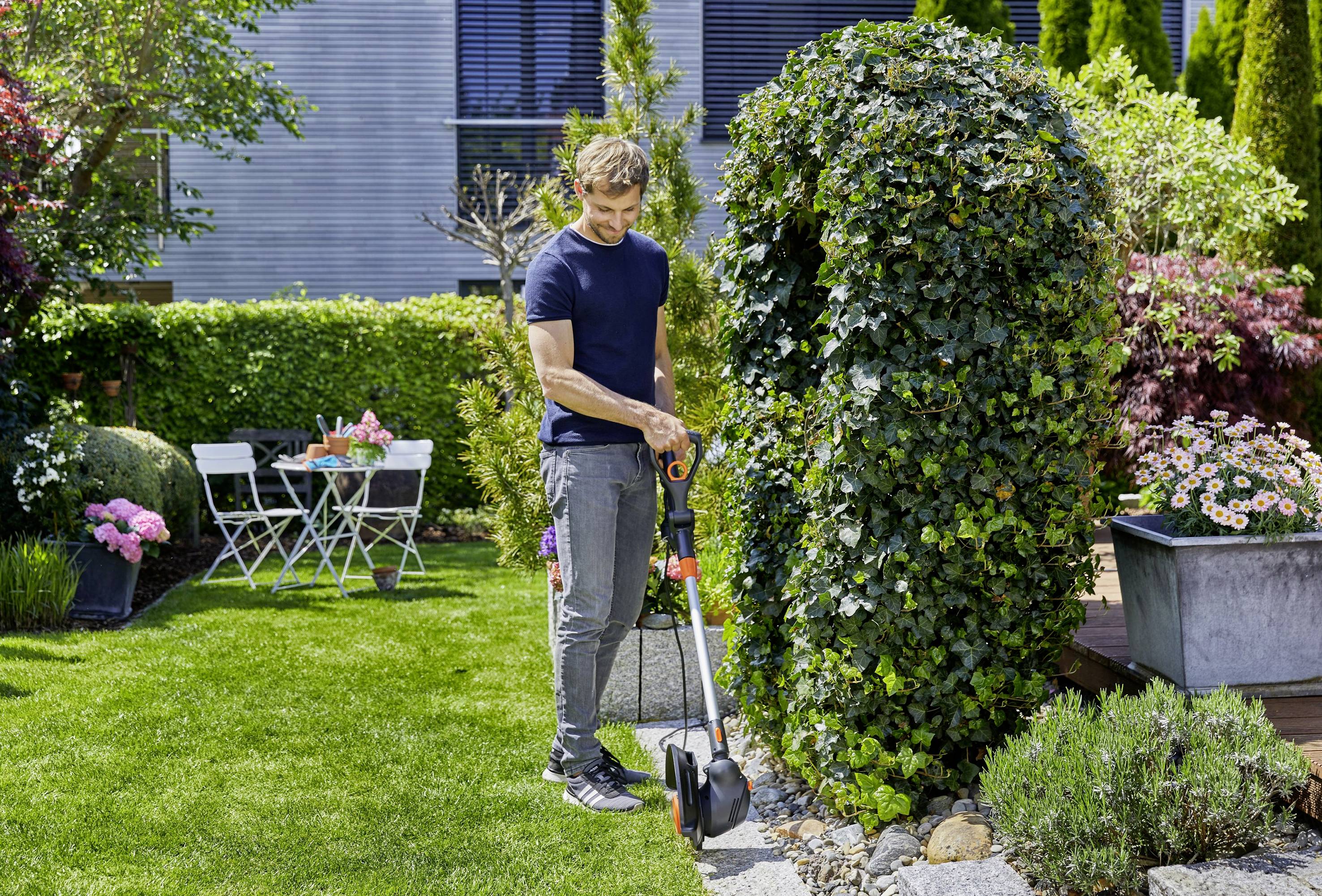 Eine Person im Garten benutzt einen Rasenkantenschneider neben einer mit Efeu bewachsenen Hecke. Im Hintergrund stehen Gartenmöbel.