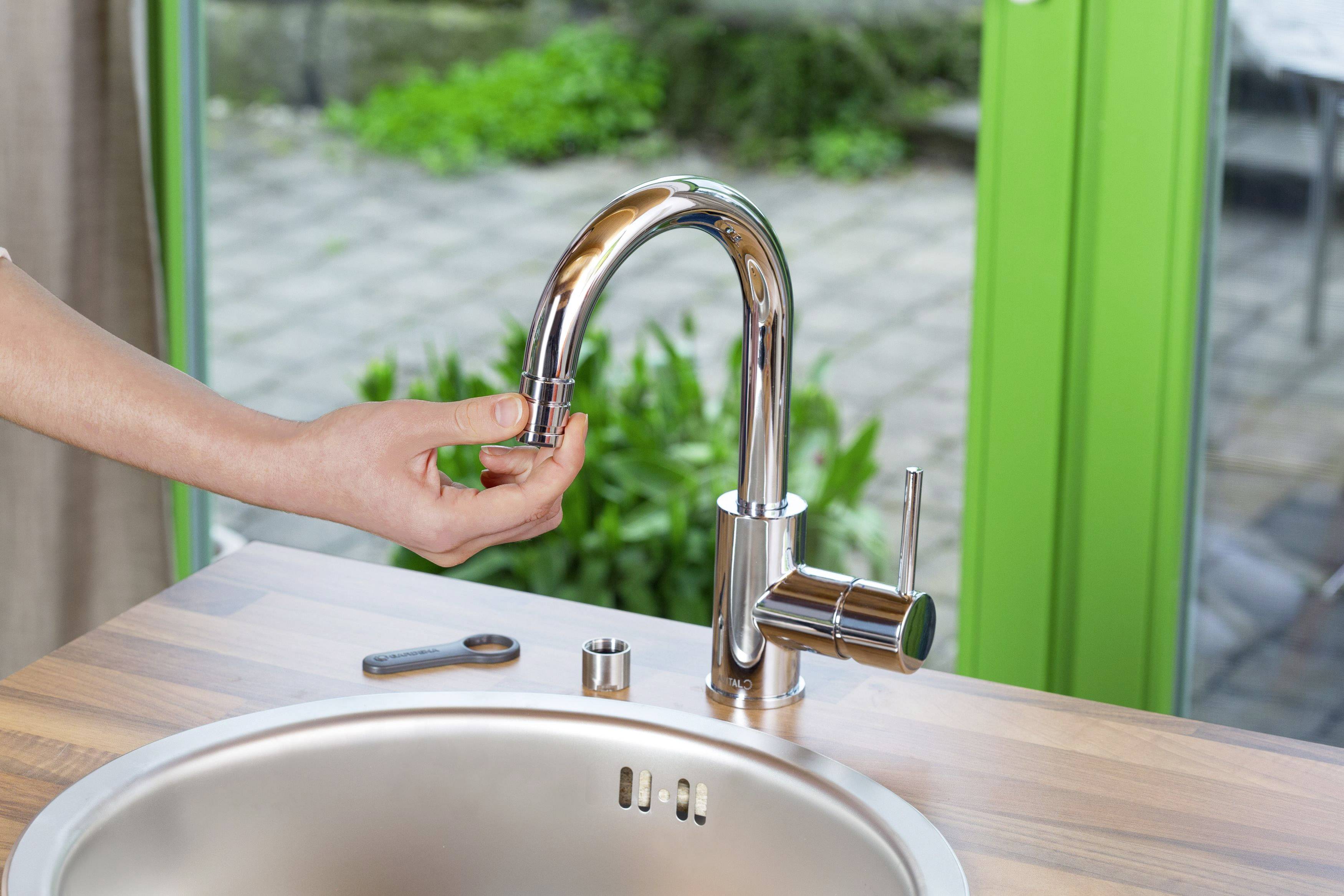 Eine Hand dreht den Strahlregler eines Küchenwasserhahns an einer Spüle locker, während im Hintergrund ein grüner Garten zu sehen ist.