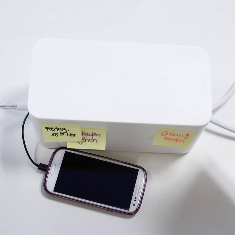 Eine weiße Kabelbox mit zwei Klebezetteln: 'Freitag, 18 Uhr, einkaufen gehen' und 'Jacken anziehen'. Ein Smartphone ist angeschlossen.