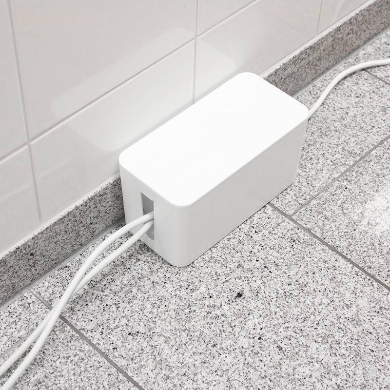 Ein weißer Kabelorganisator aus Kunststoff steht auf einem gefliesten Boden an der Wand. Zwei Kabel treten links heraus.