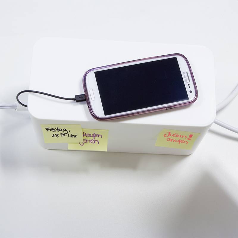 Ein Smartphone wird aufgeladen, liegt auf einer weißen Box. Zwei Notizzettel: 'Freitag 18-22 Uhr kaufen gehen' und 'Schulen anrufen'.