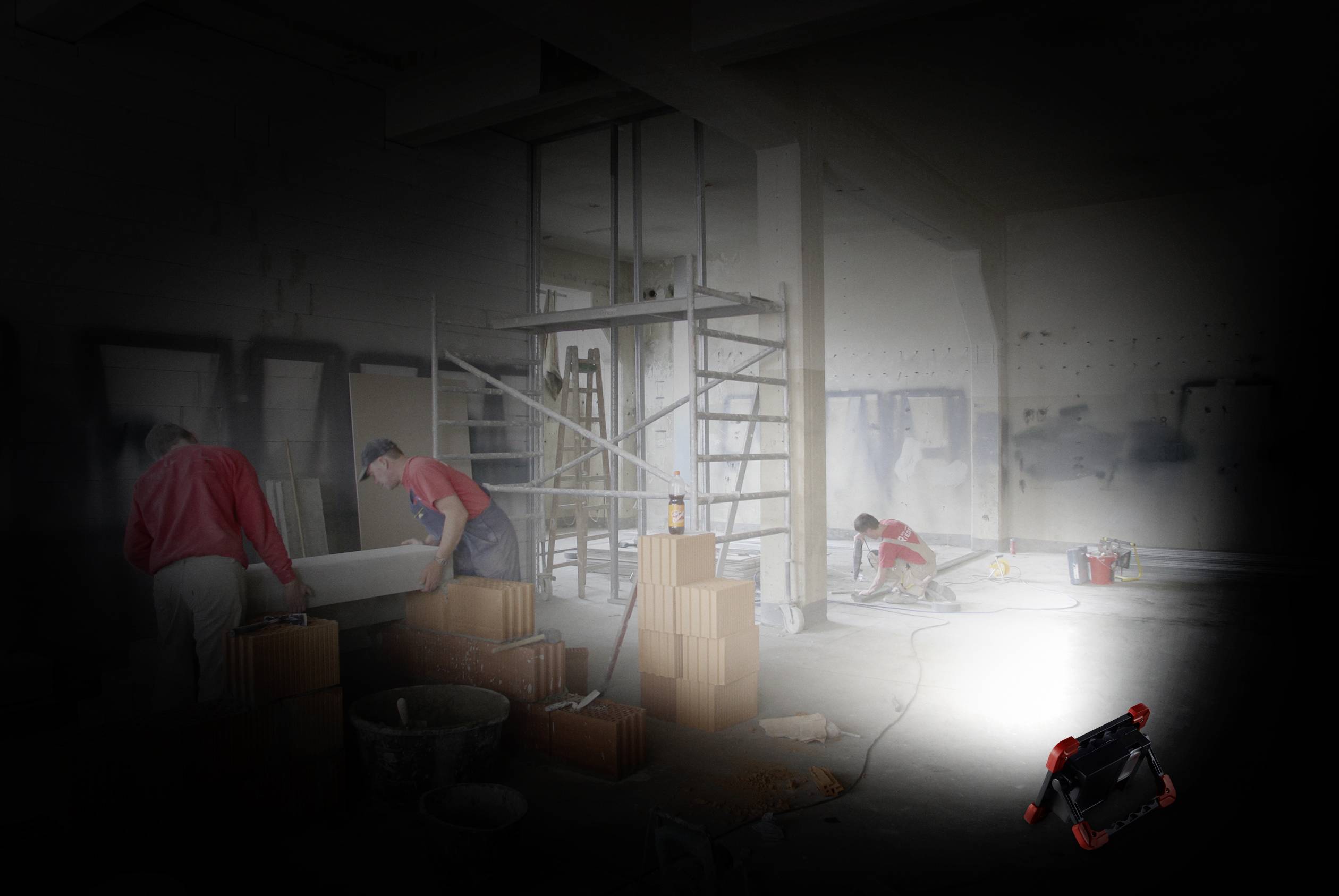 Bauarbeiter arbeiten auf einer Baustelle. Einer trägt Ziegel, ein anderer kniet, um Kabel zu verlegen. Eine Lichtquelle beleuchtet den Raum.