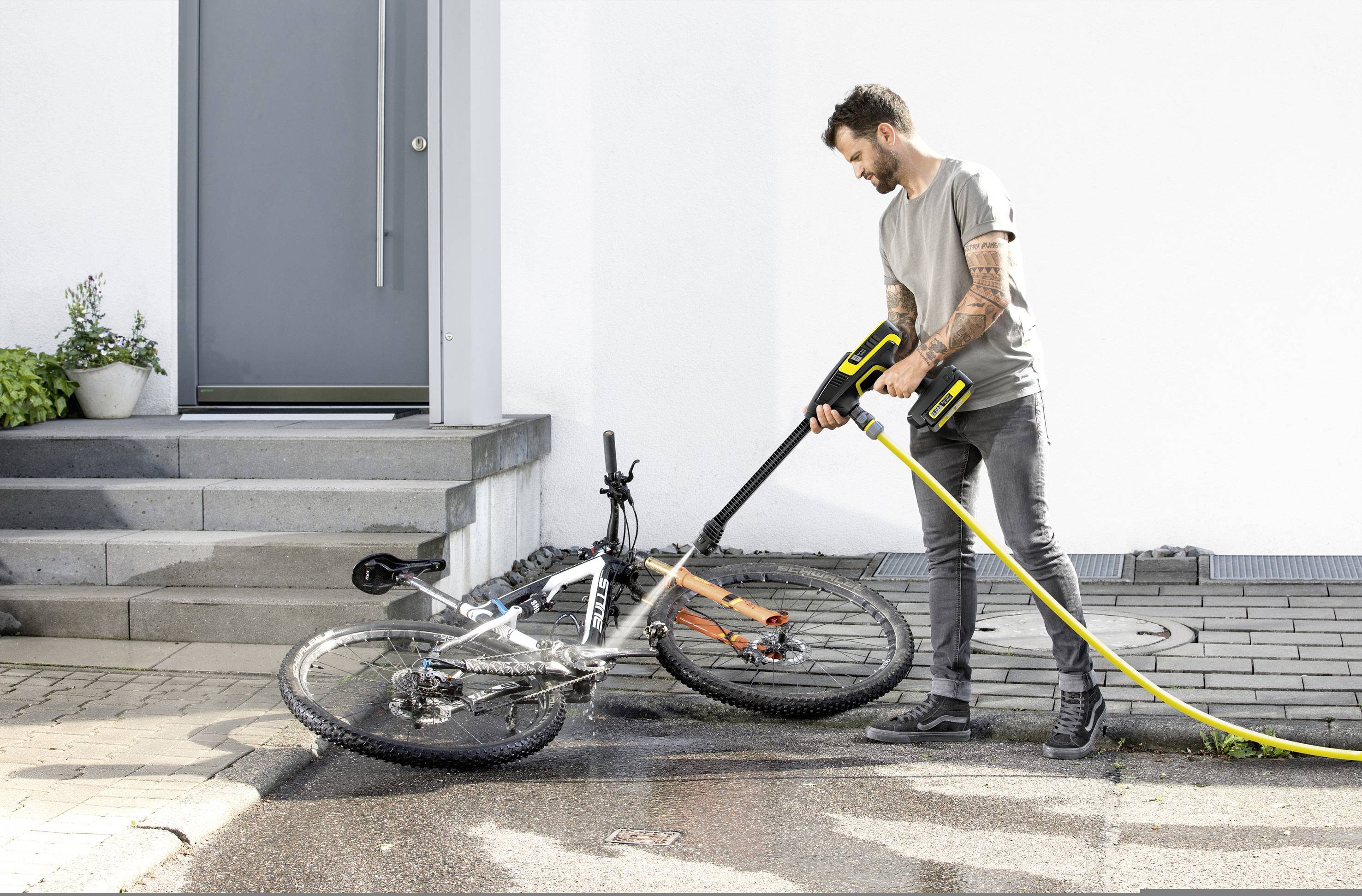 Ein Mann reinigt ein Fahrrad mit einem Hochdruckreiniger vor einem Hauseingang.