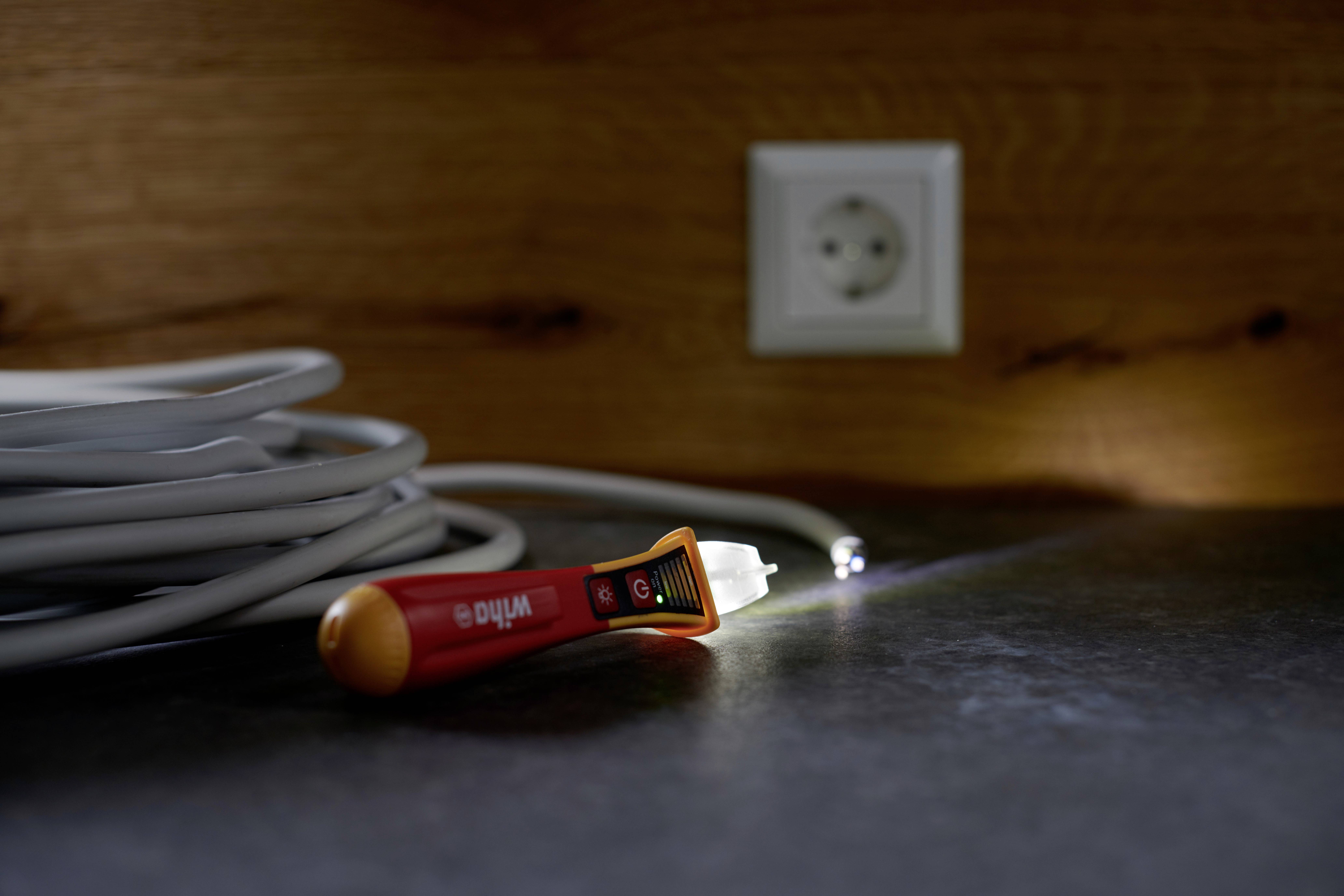 Ein zusammengerolltes weißes Kabel liegt auf einer Arbeitsfläche. Im Hintergrund ist eine Steckdose. Ein Spannungsprüfer leuchtet darauf.