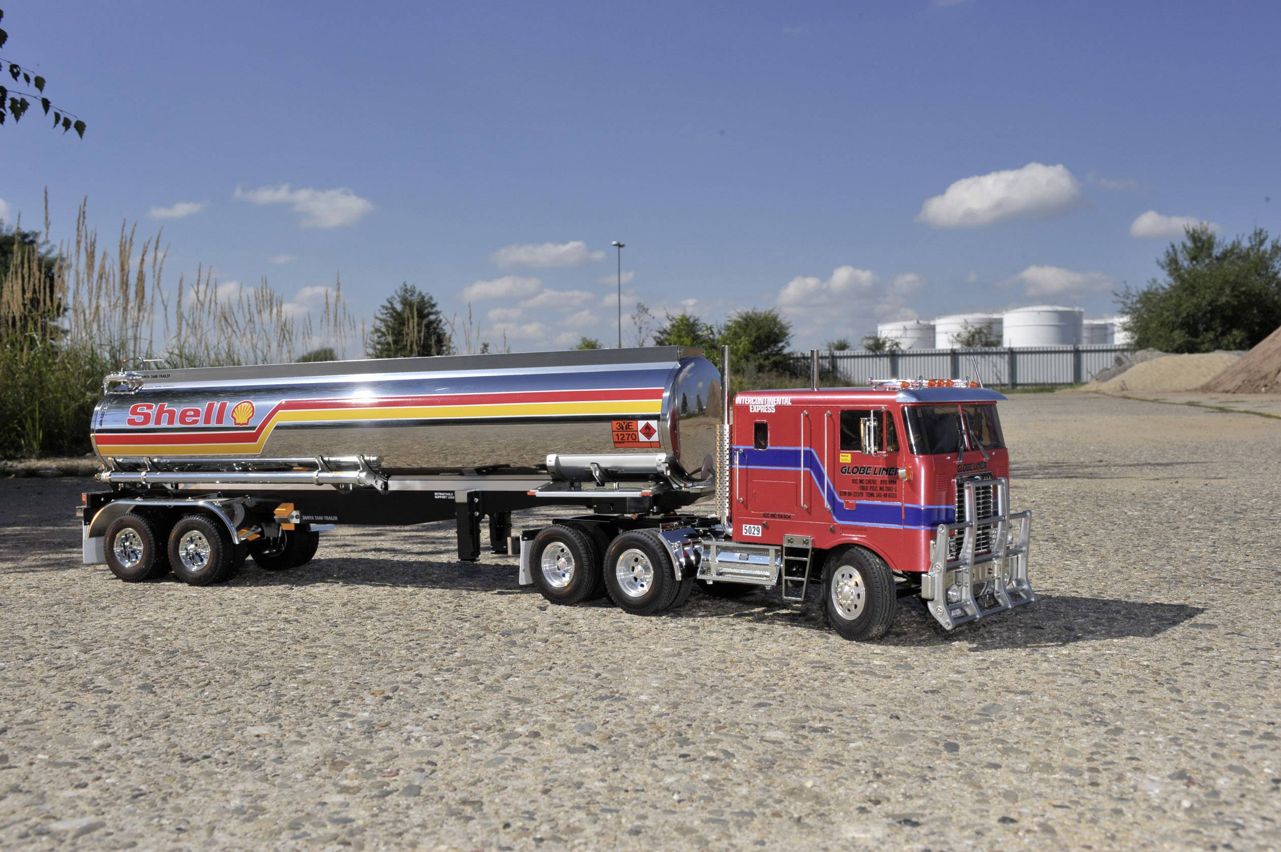 Ein roter Tanklastwagen mit dem Shell-Logo steht auf einem Kiesweg. Im Hintergrund sind Bäume und Industriebauten zu sehen.