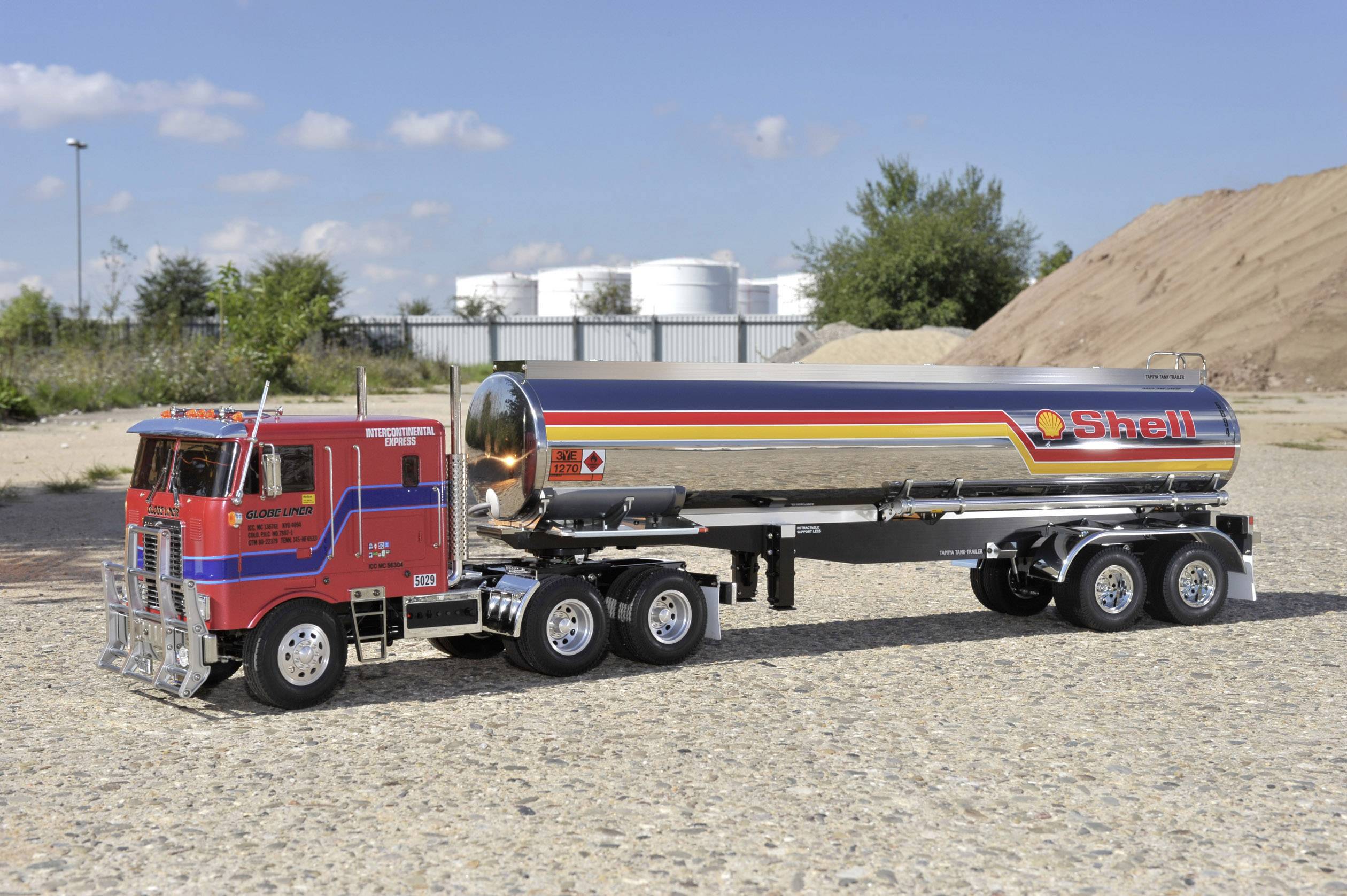 Ein Modell-LKW mit Tankauflieger im Shell-Design steht auf einem Betonplatz. Hintergrund zeigt Lagergebäude und Sandhaufen.