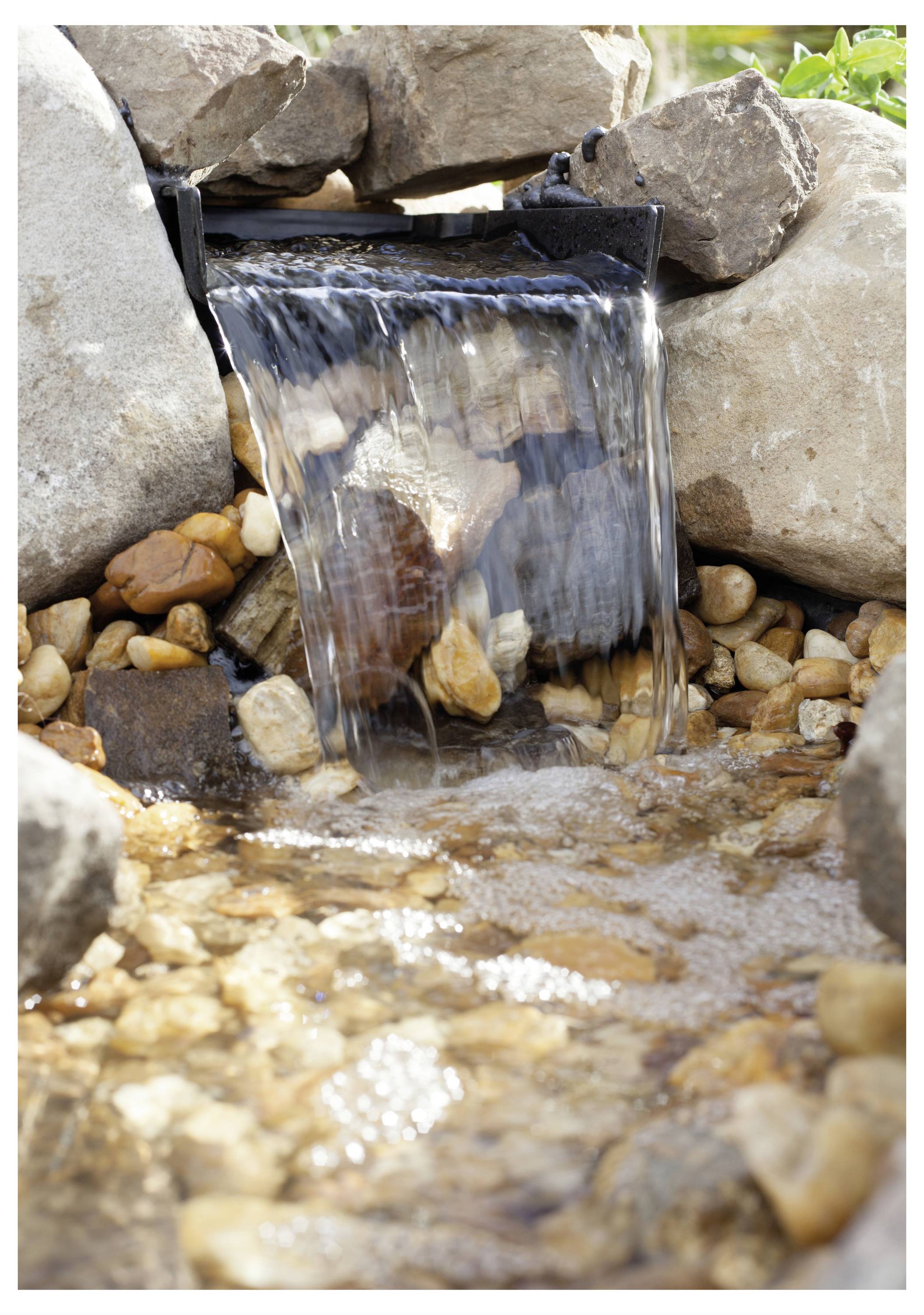 Ein kleiner Wasserfall fließt über Felsen in einen seichten Bach, umgeben von größeren Steinen und Kieseln, in einer natürlichen Gartenlandschaft.