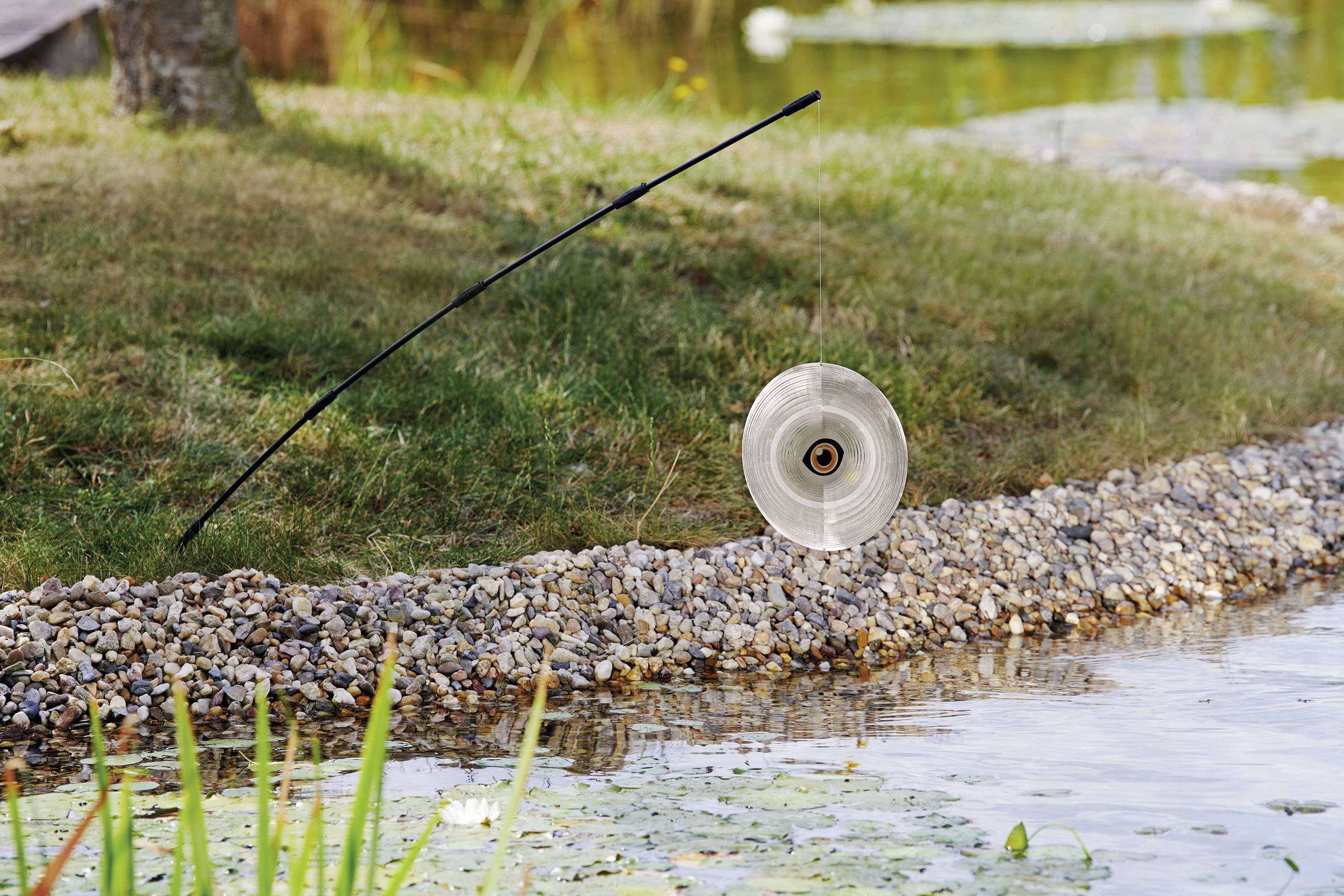 Ein stehender Gartenschlauchhalter mit einem aufgerollten Schlauch auf einem steinigen Ufer neben einem Teich mit Seerosen.