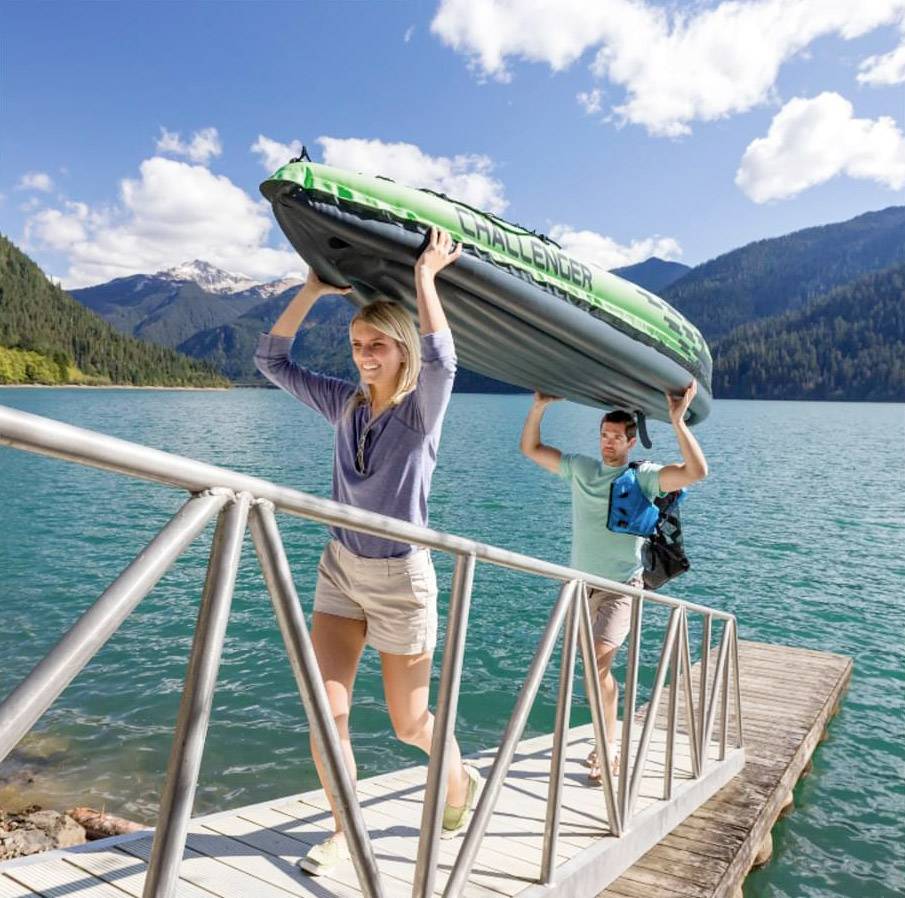 Zwei Personen tragen gemeinsam ein aufblasbares Kajak über einen Steg an einem Bergsee bei sonnigem Wetter. Berge und Wolken im Hintergrund.