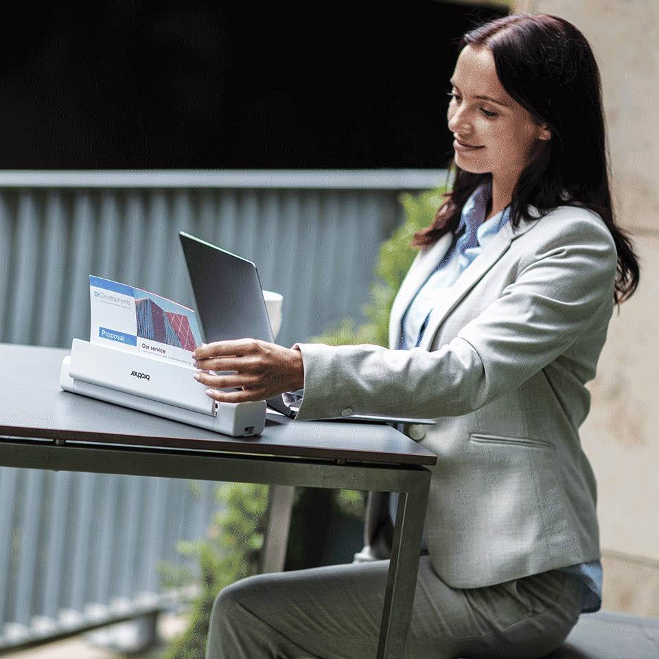 Eine Frau in einem grauen Anzug sitzt draußen an einem Tisch und nutzt einen Scanner. Neben ihr steht ein Laptop. Im Hintergrund sind Pflanzen zu sehen.