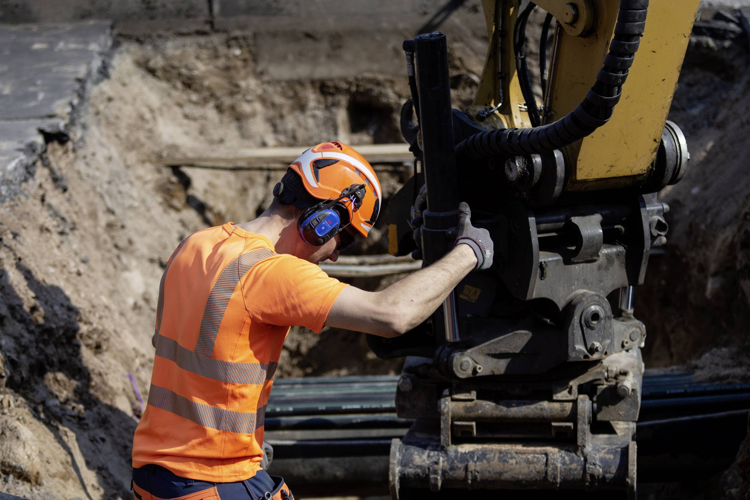 Ein Bauarbeiter in Schutzkleidung bedient einen Bagger auf einer Baustelle. Er trägt einen Helm und Gehörschutz, während er arbeitet.