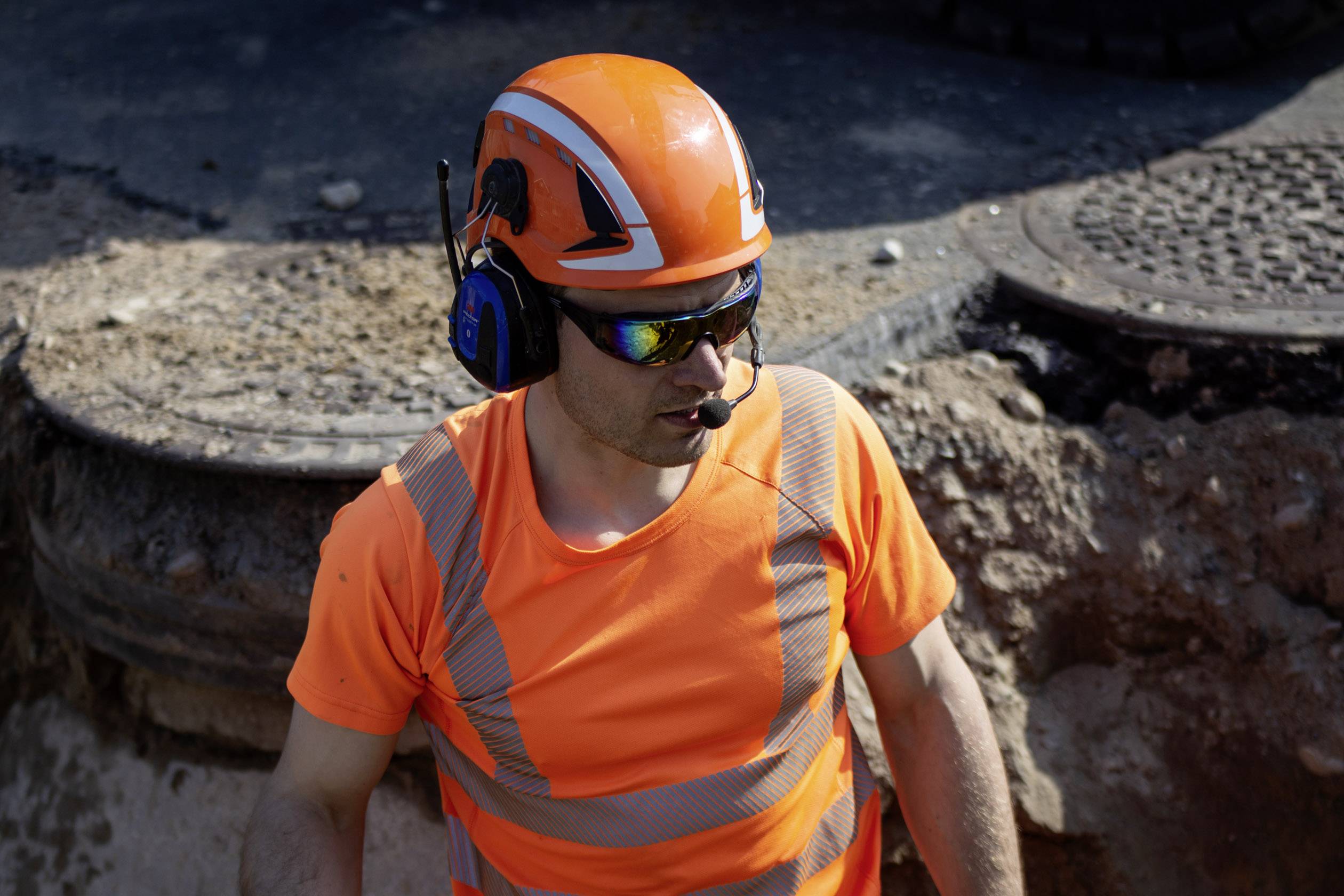 Bauarbeiter mit orangem Helm und Schutzkleidung arbeitet auf einer Baustelle. Er trägt Kopfhörer mit Mikrofon und reflektierende Weste.