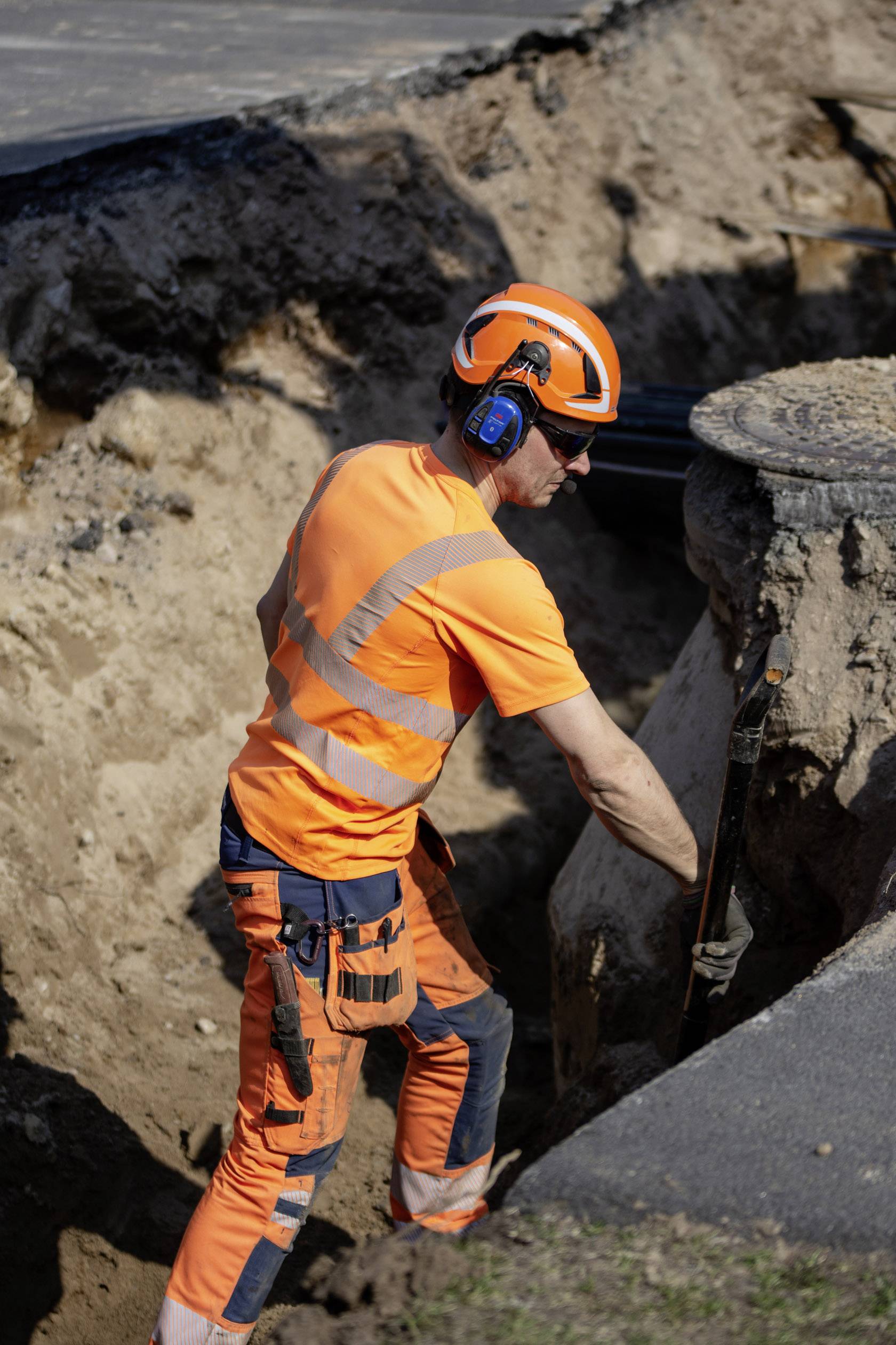 Ein Bauarbeiter in orangefarbener Schutzkleidung arbeitet in einer tiefen Grube auf einer Baustelle. Er trägt Helm und Gehörschutz.