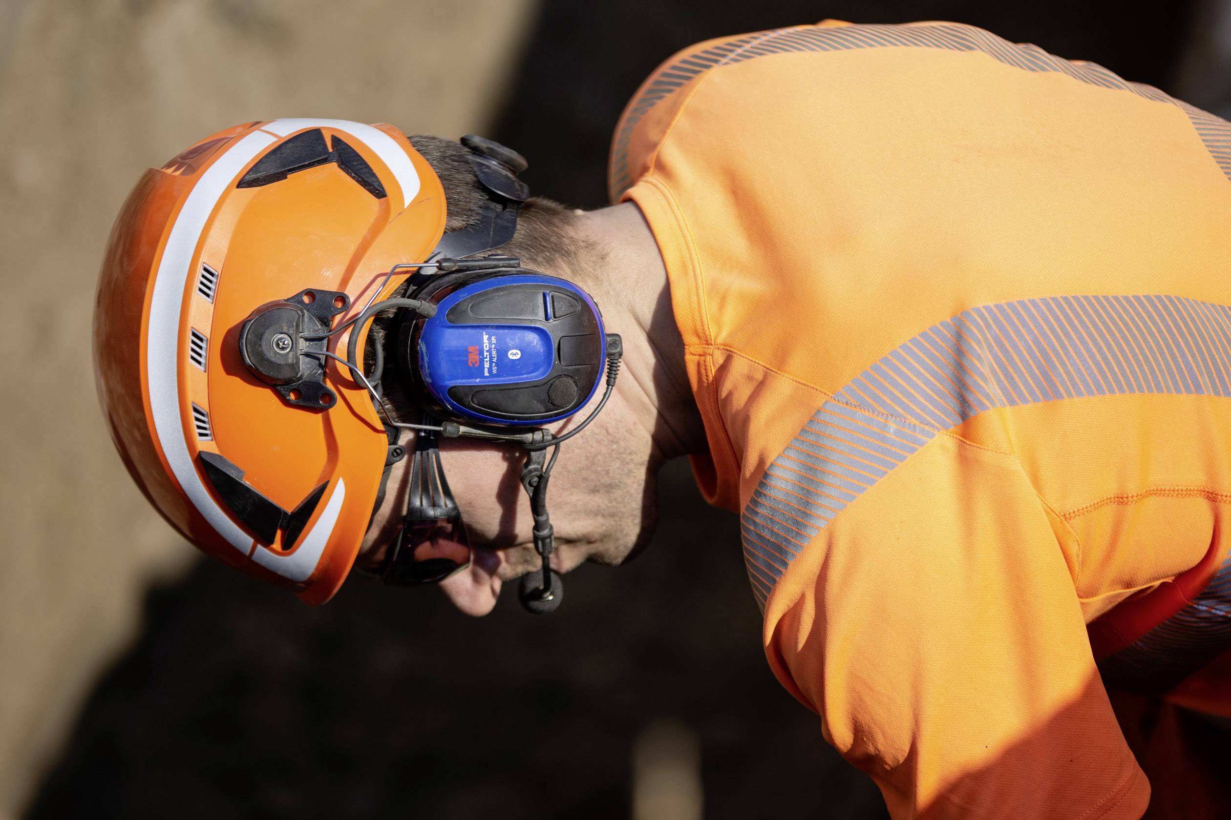 Ein Bauarbeiter mit orangefarbener Schutzkleidung und Helm, trägt Gehörschutz mit Mikrofon. Fokussiert auf seine Arbeit.