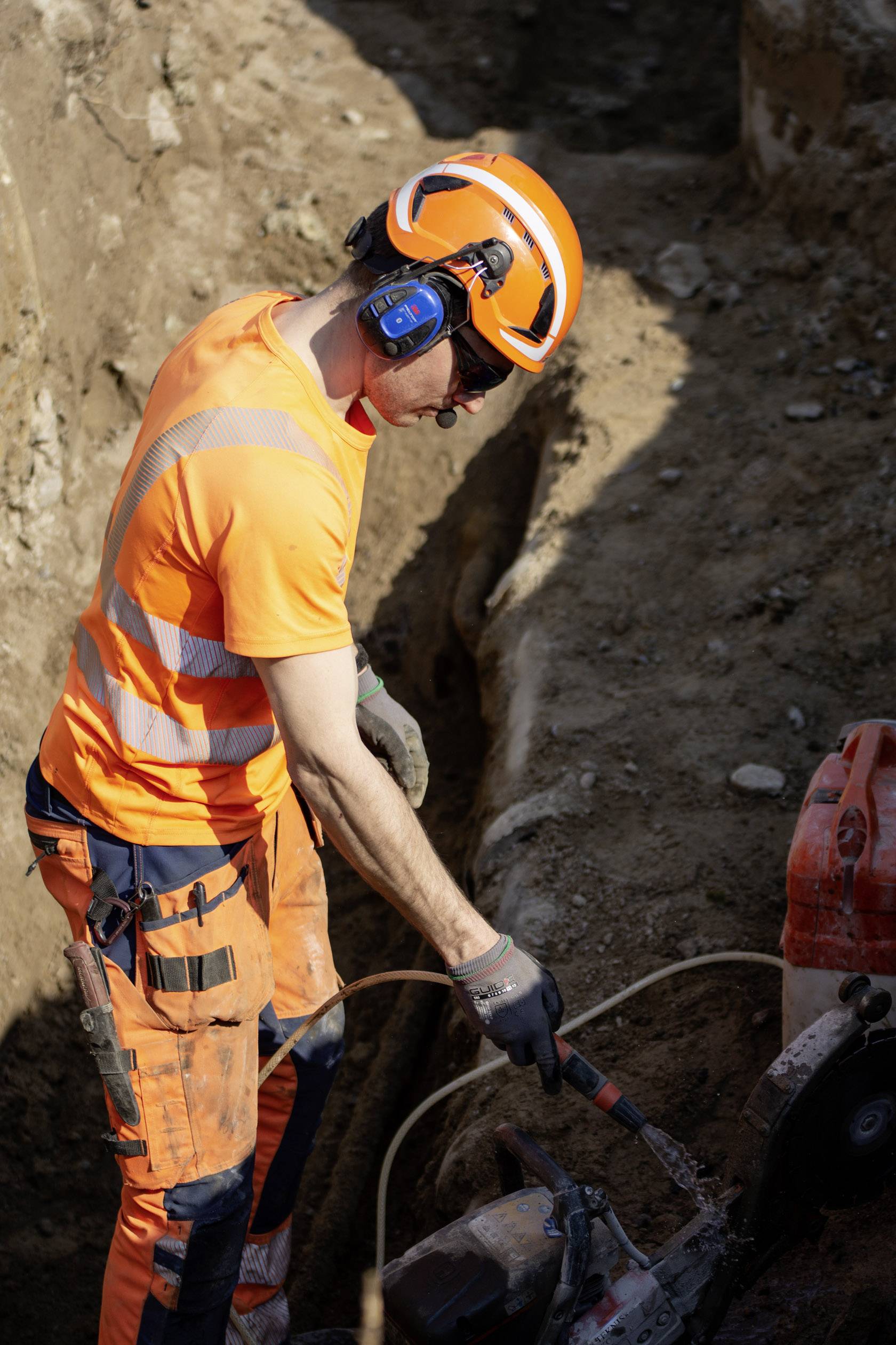 Eine Person arbeitet auf einer Baustelle, trägt orangene Sicherheitskleidung und einen Helm. Der Ort ist staubig mit Sand und Erde.